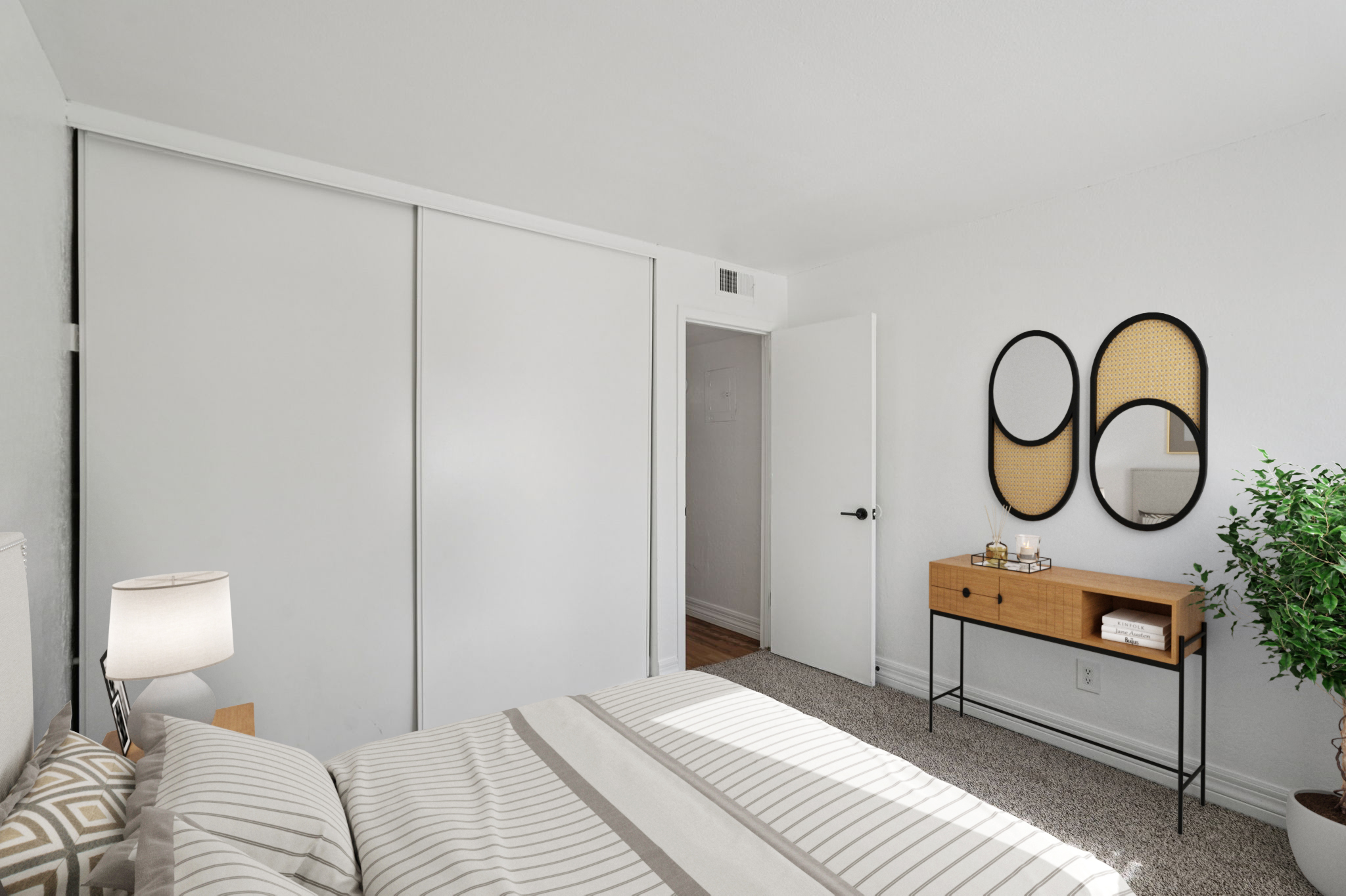 A bright, modern bedroom featuring a bed with a striped comforter, a bedside lamp, and a dresser with decorative items. The walls are white, and there are two round mirrors above the dresser. A potted plant adds a touch of greenery, and a closet with sliding doors is visible in the background.