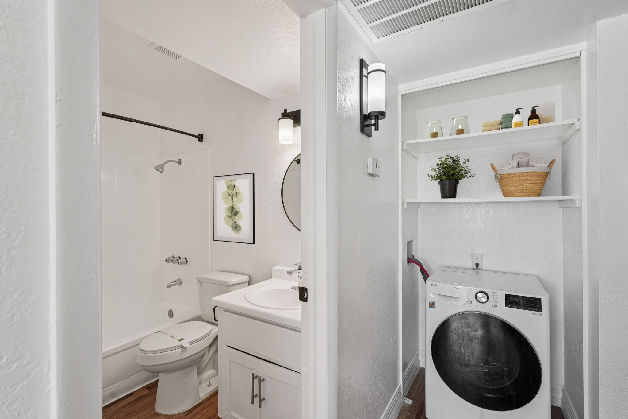 A modern bathroom featuring a bathtub with a shower, a white toilet, and a sink with a mirror. Adjacent to the bathroom is a laundry area with a white washing machine, shelves displaying toiletries and a plant, and bright lighting, creating a clean and functional space.