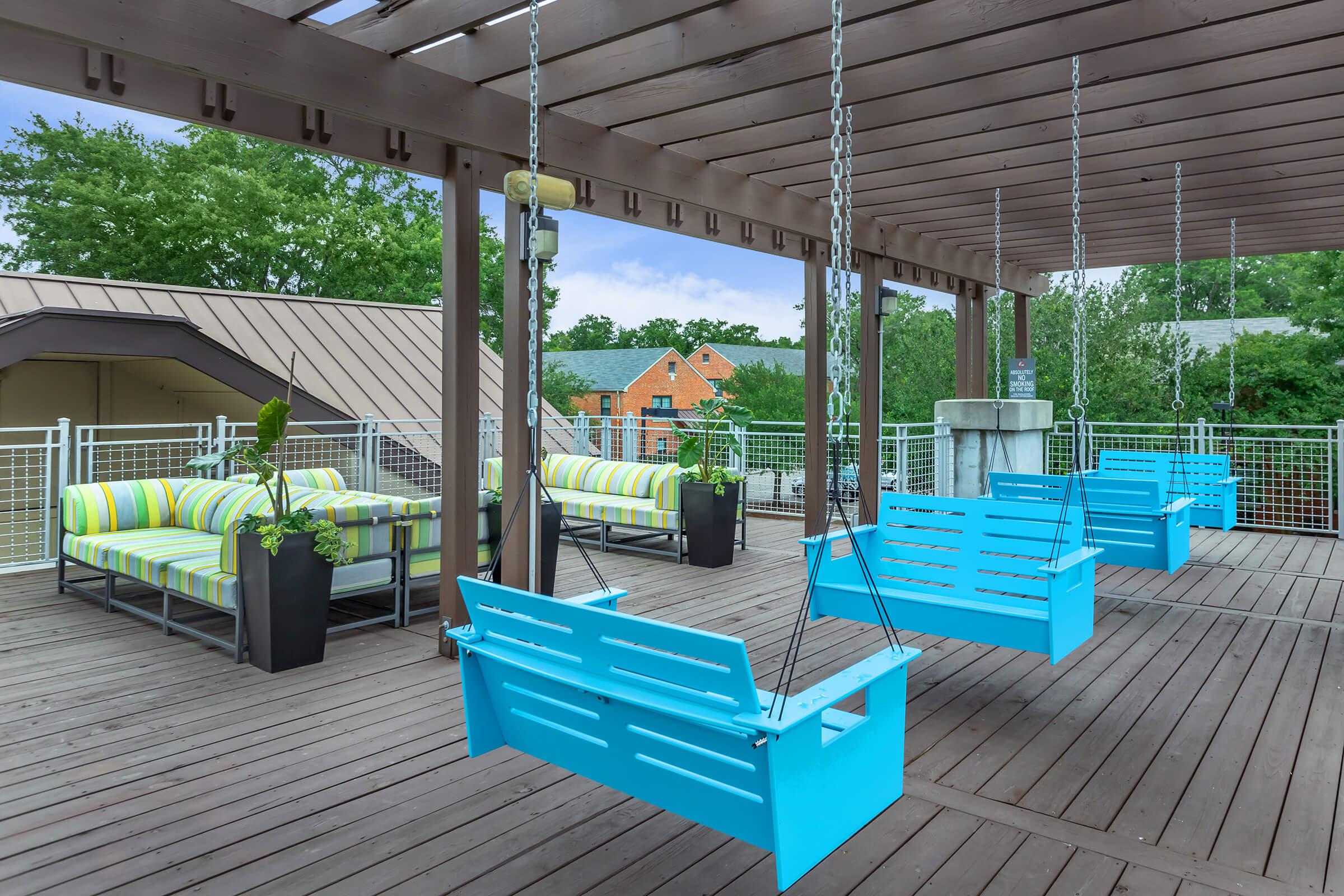 A modern outdoor patio with wooden flooring, featuring several turquoise swing chairs and brightly striped sofas. The area is surrounded by greenery and includes potted plants, creating a relaxing atmosphere for socializing or enjoying the outdoors. The clear blue sky complements the inviting space.