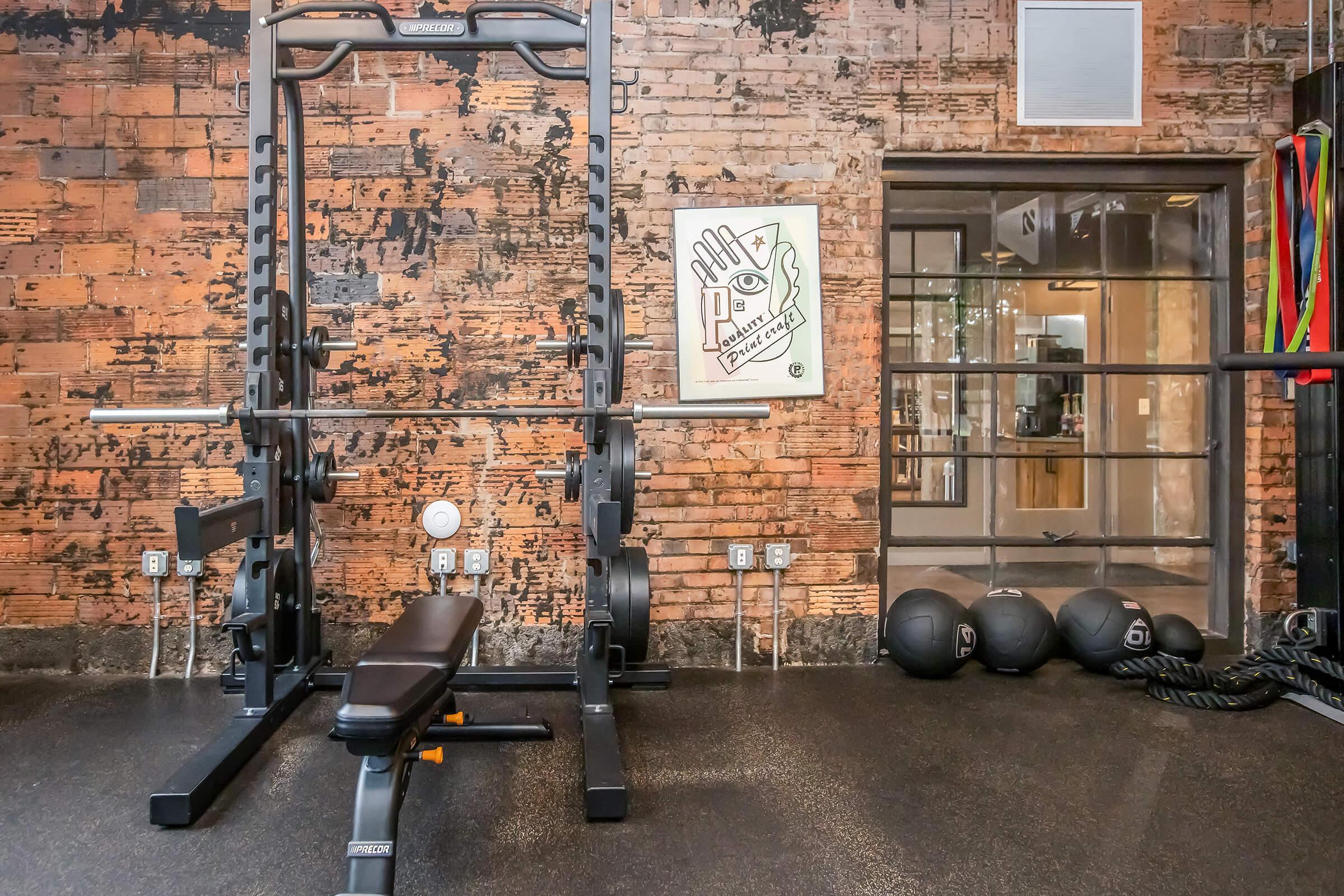 A modern gym interior featuring a weightlifting squat rack, a flat bench, and several black medicine balls on the floor. The wall has a rustic brick finish, and there is a motivational poster visible in the background, along with various fitness bands hanging on the side.