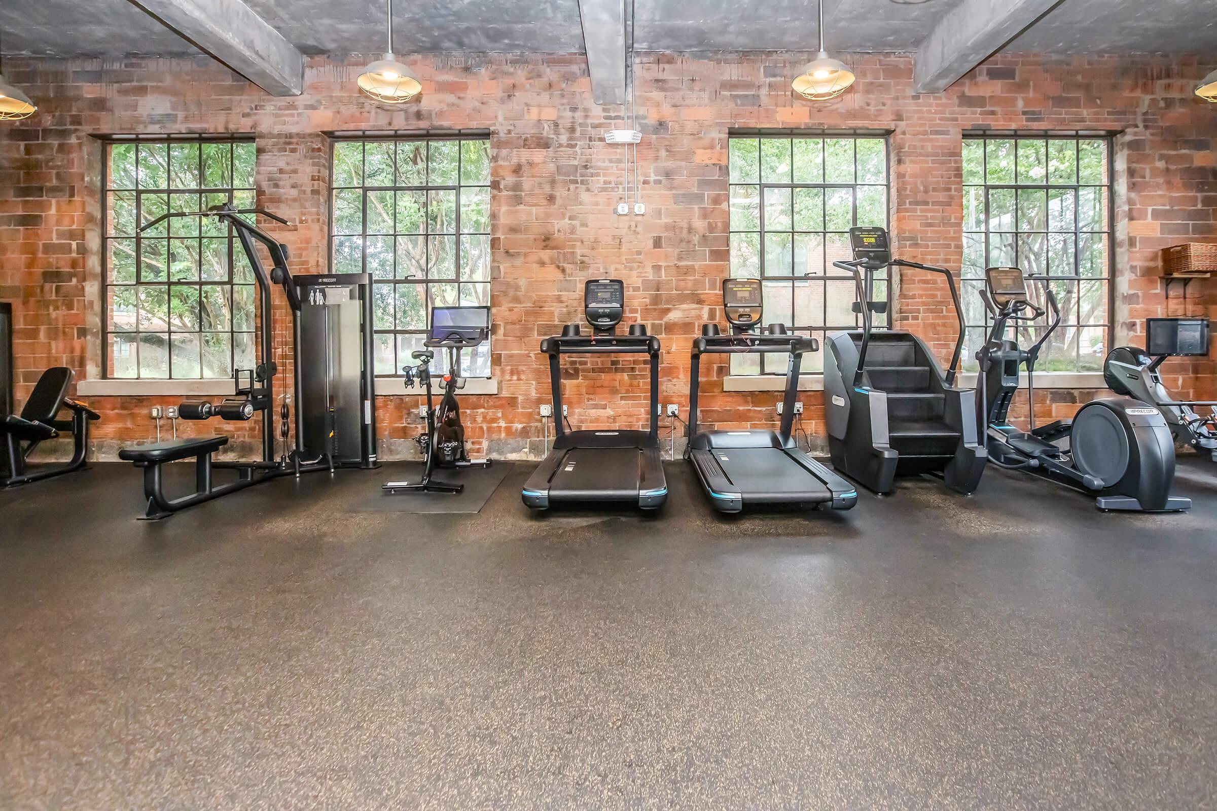 A modern gym space featuring various fitness equipment, including treadmills, an elliptical machine, a stationary bike, and strength training equipment. The room has industrial-style brick walls and large windows allowing natural light to illuminate the area, creating an inviting atmosphere for workouts.