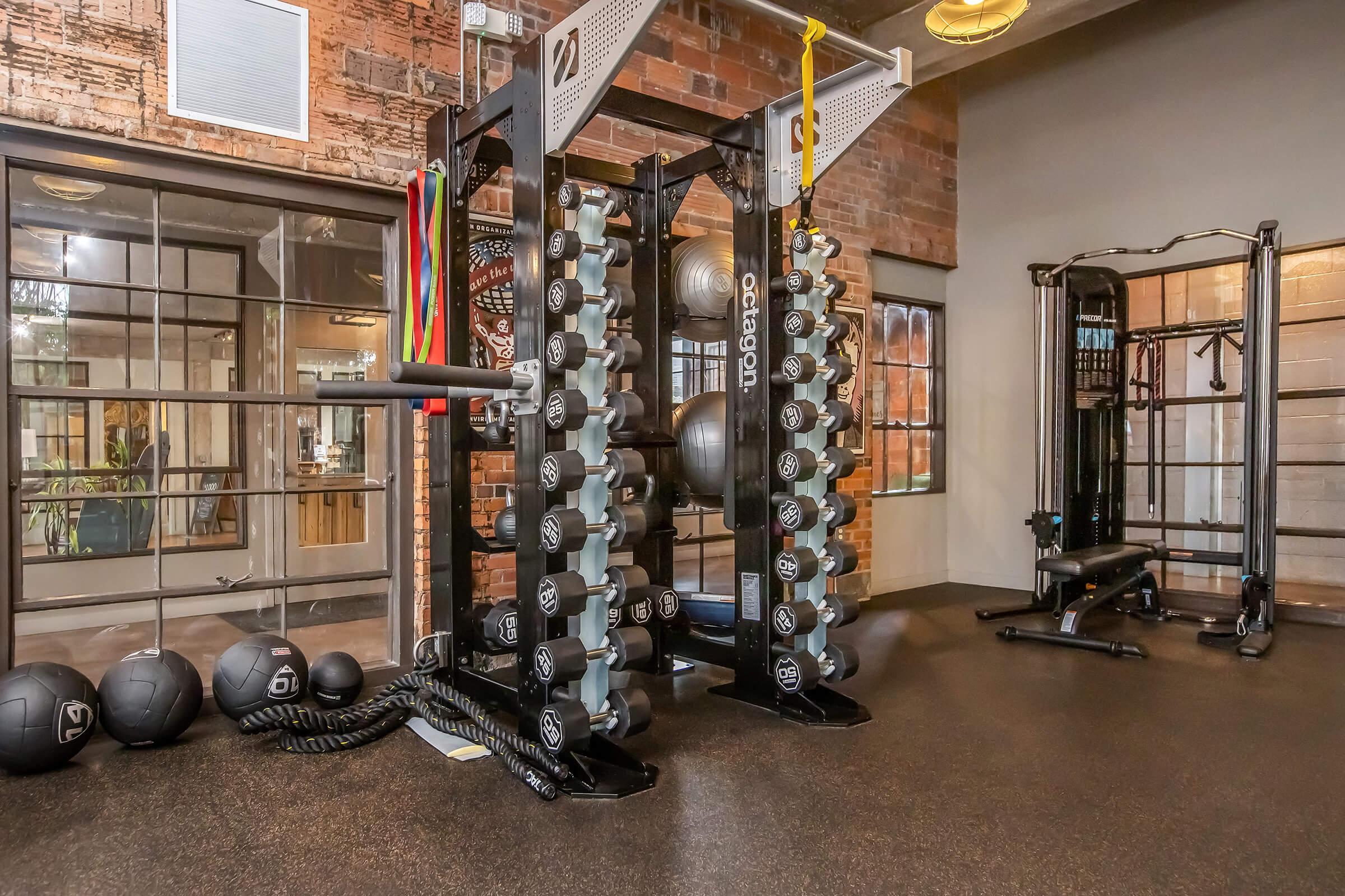 A modern gym interior featuring a weight rack with various dumbbells, stability balls, and exercise equipment. The space has a brick wall backdrop and large windows, contributing to a bright and inviting atmosphere for workouts.