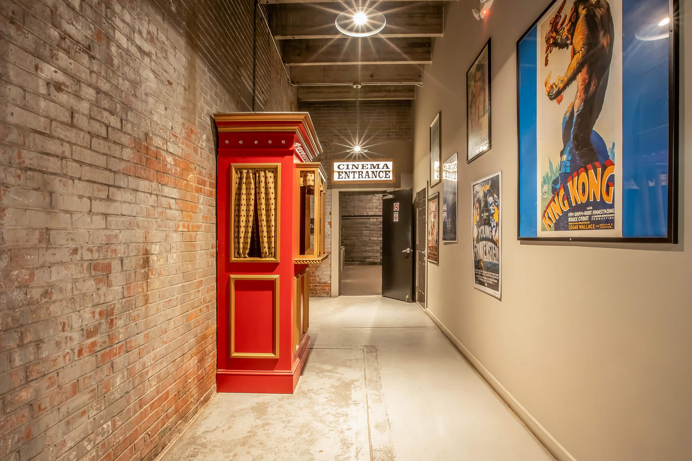 A corridor leading to a cinema entrance. The walls feature exposed brick and posters of classic movies. A vintage-style ticket booth in red stands on the left, while a door at the end indicates the cinema entrance. Soft overhead lighting enhances the inviting atmosphere.