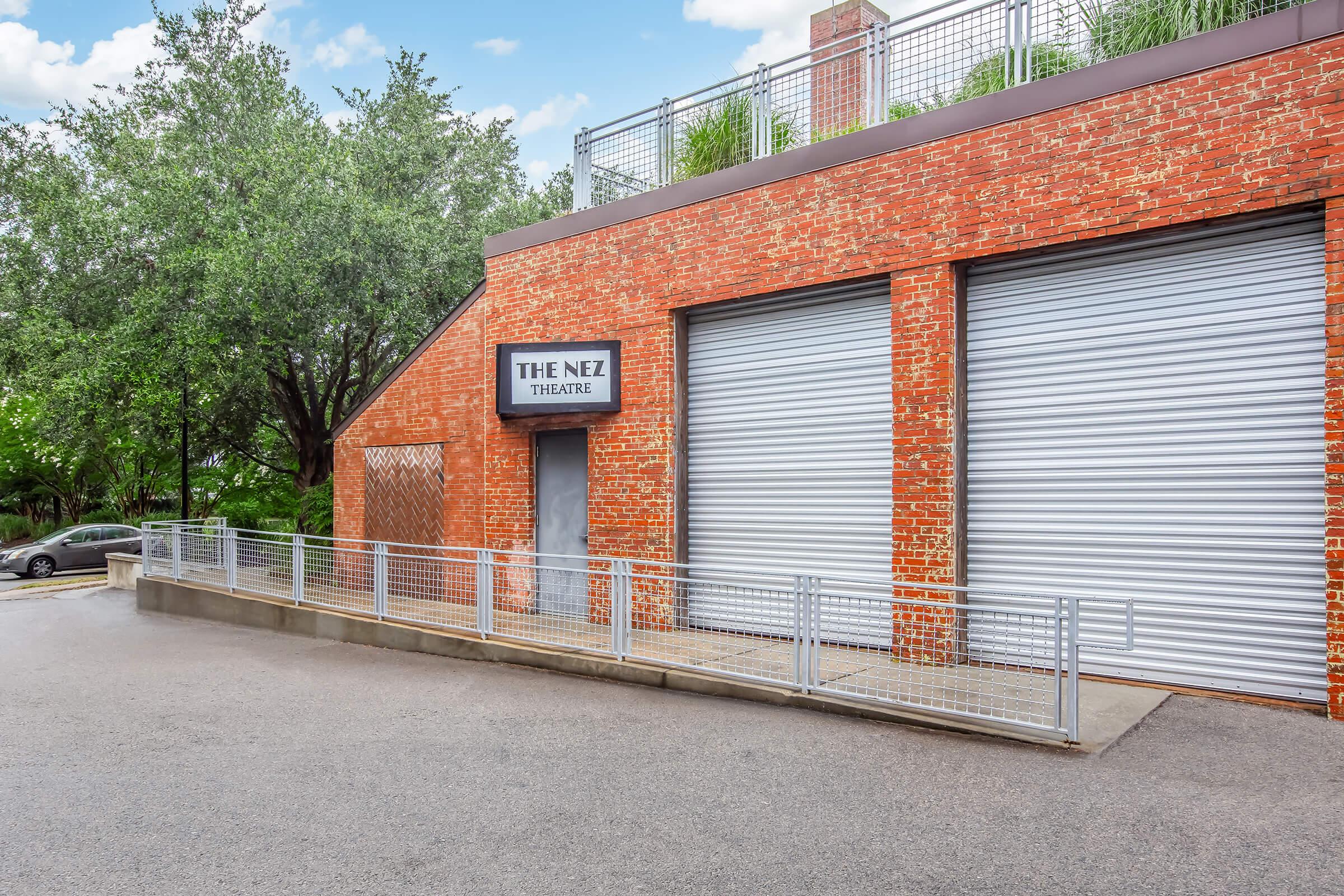 Exterior view of The Nez Theatre, featuring a brick facade with large gray garage doors, a sign displaying "THE NEZ THEATRE," and a ramp leading up to the entrance. Surrounding the building are green trees and a clear sky. The area appears clean and well-maintained.