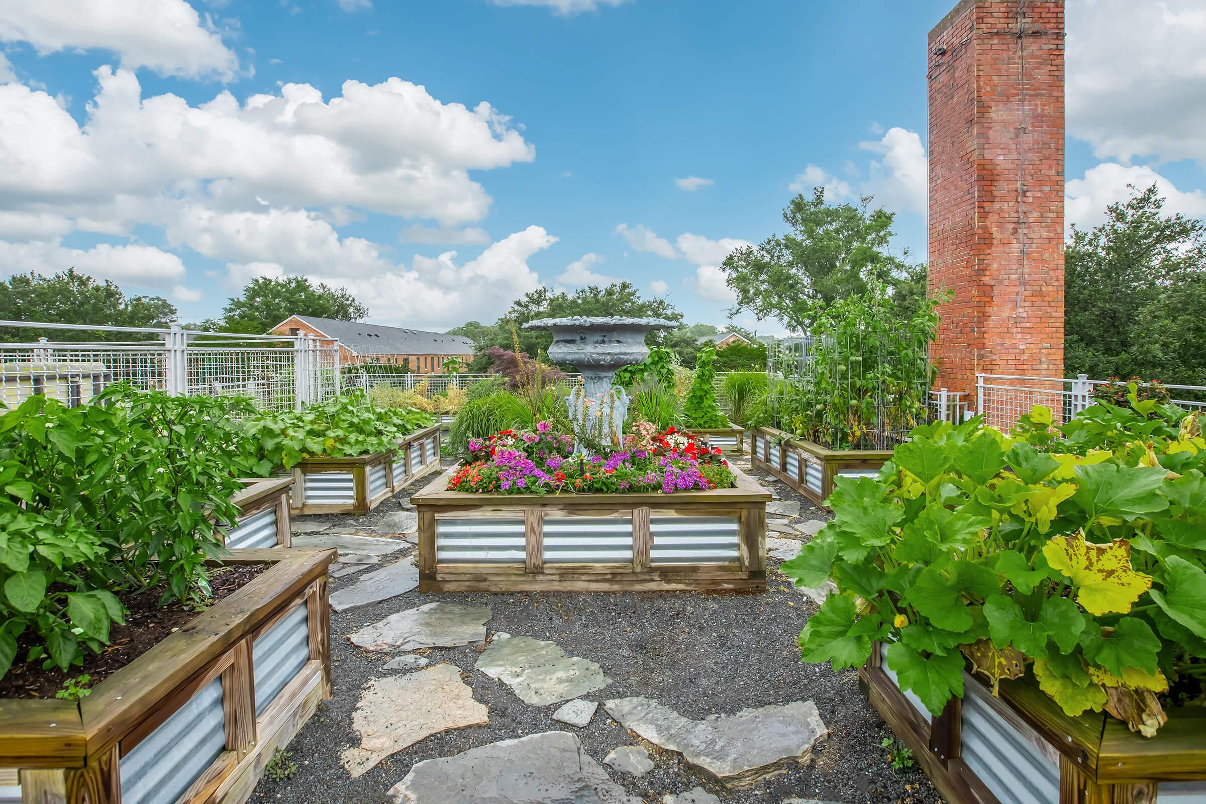A rooftop garden featuring rectangular raised planting beds filled with various vegetables and vibrant flowers. In the background, there are trees and a brick chimney. The sky is partly cloudy, adding a bright and open feel to the scene.