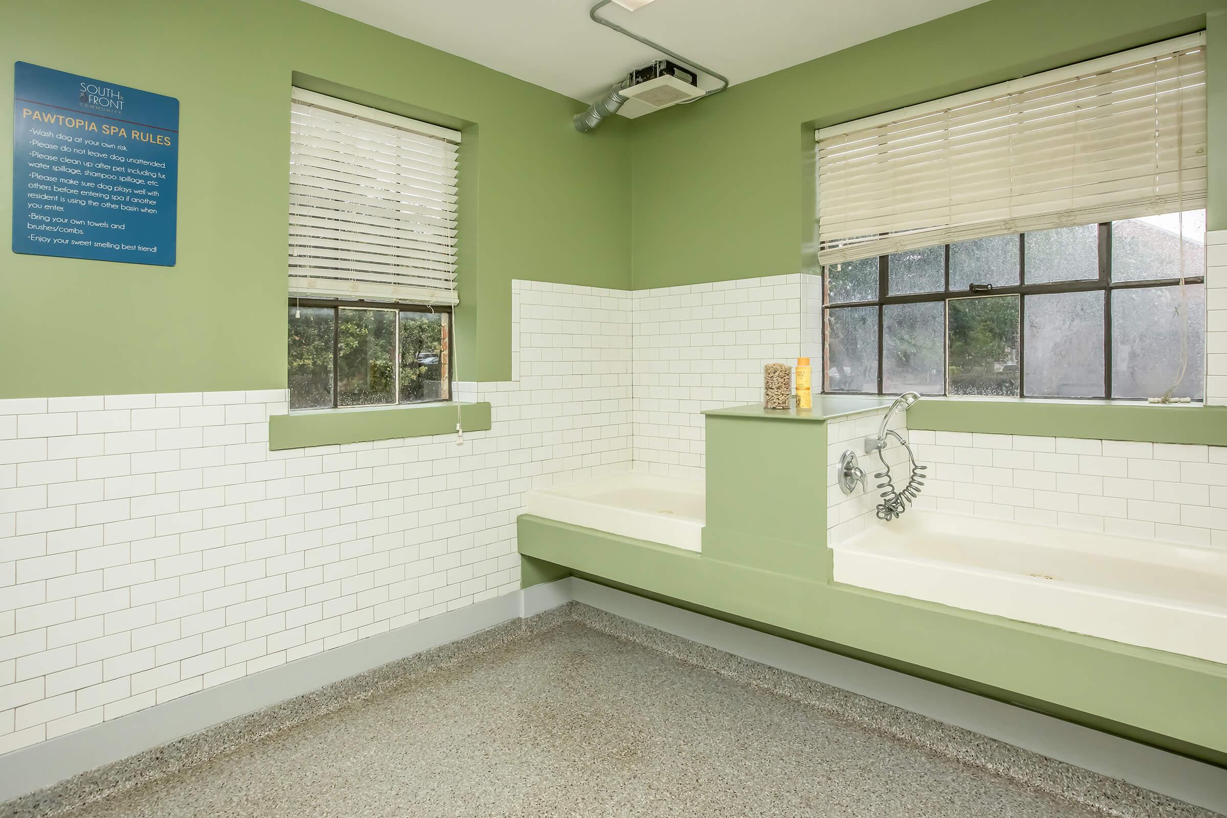 A bright room with green walls and white tiled areas, featuring two bathtubs. There's a shower attachment on the wall. Large windows with blinds allow natural light. A sign on the wall outlines spa rules, and the floor is finished with a speckled light gray material.
