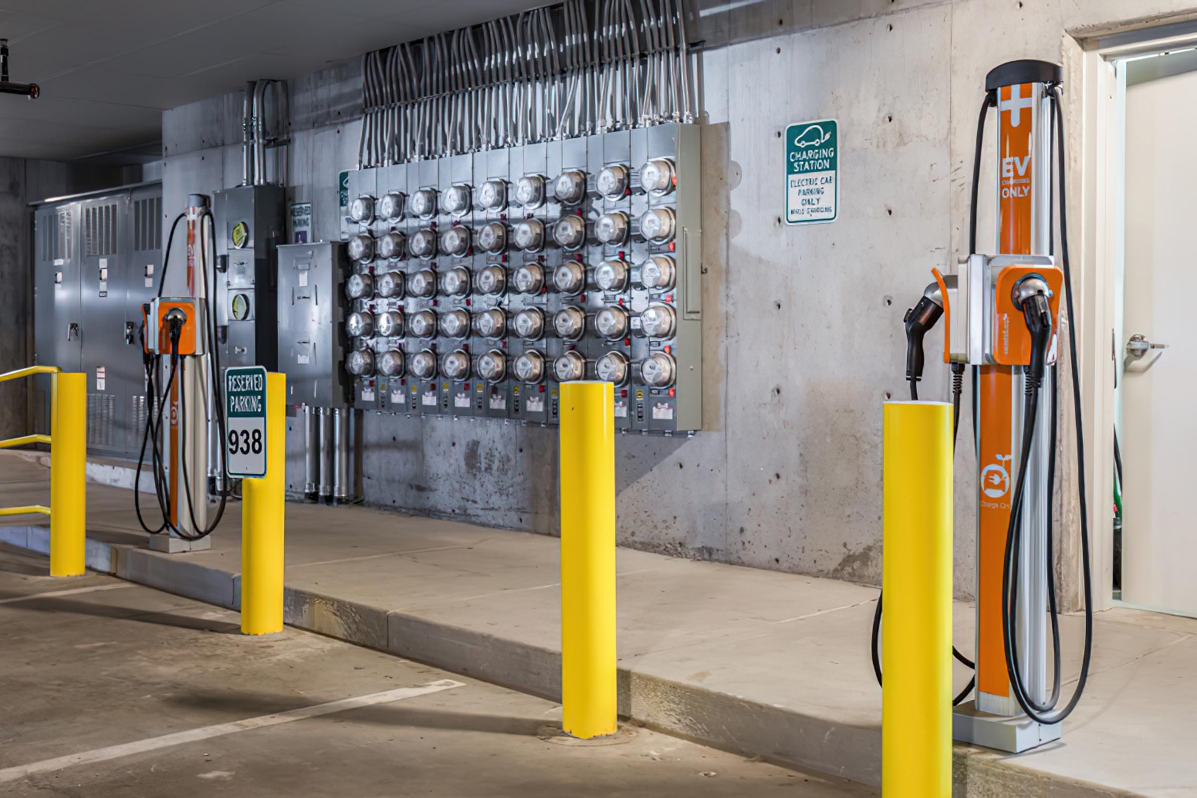 Electric vehicle charging station in a parking garage, featuring multiple charging units attached to the wall and two standalone charging posts with cables. Yellow safety posts are positioned nearby, and there are signs indicating electric vehicle parking and charging station usage.