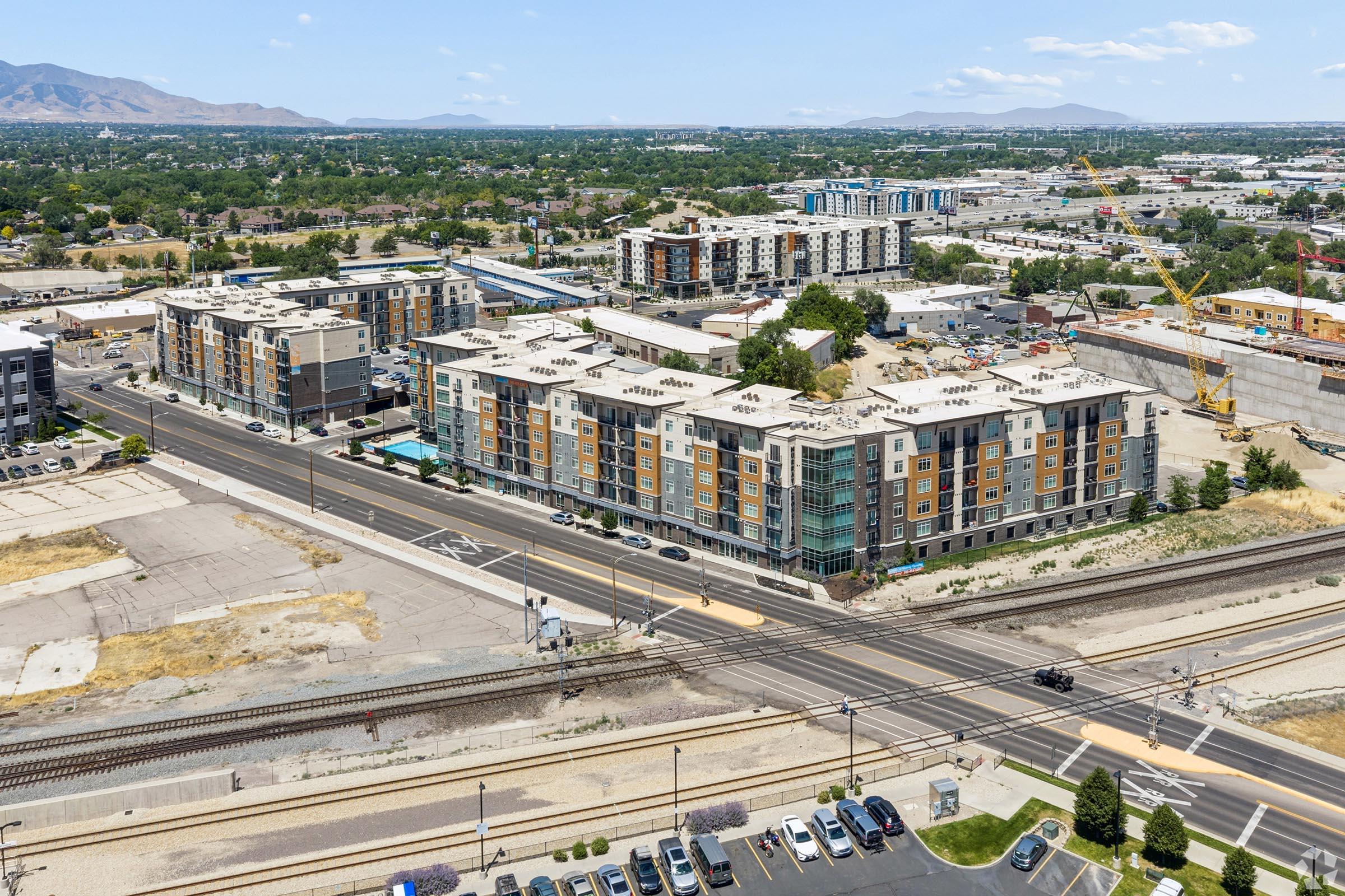 Aerial view of a multi-story residential building next to railway tracks and a road. The surrounding area includes other buildings, greenery, and distant mountains under a clear blue sky. Urban landscape shows a mix of developed and undeveloped land.