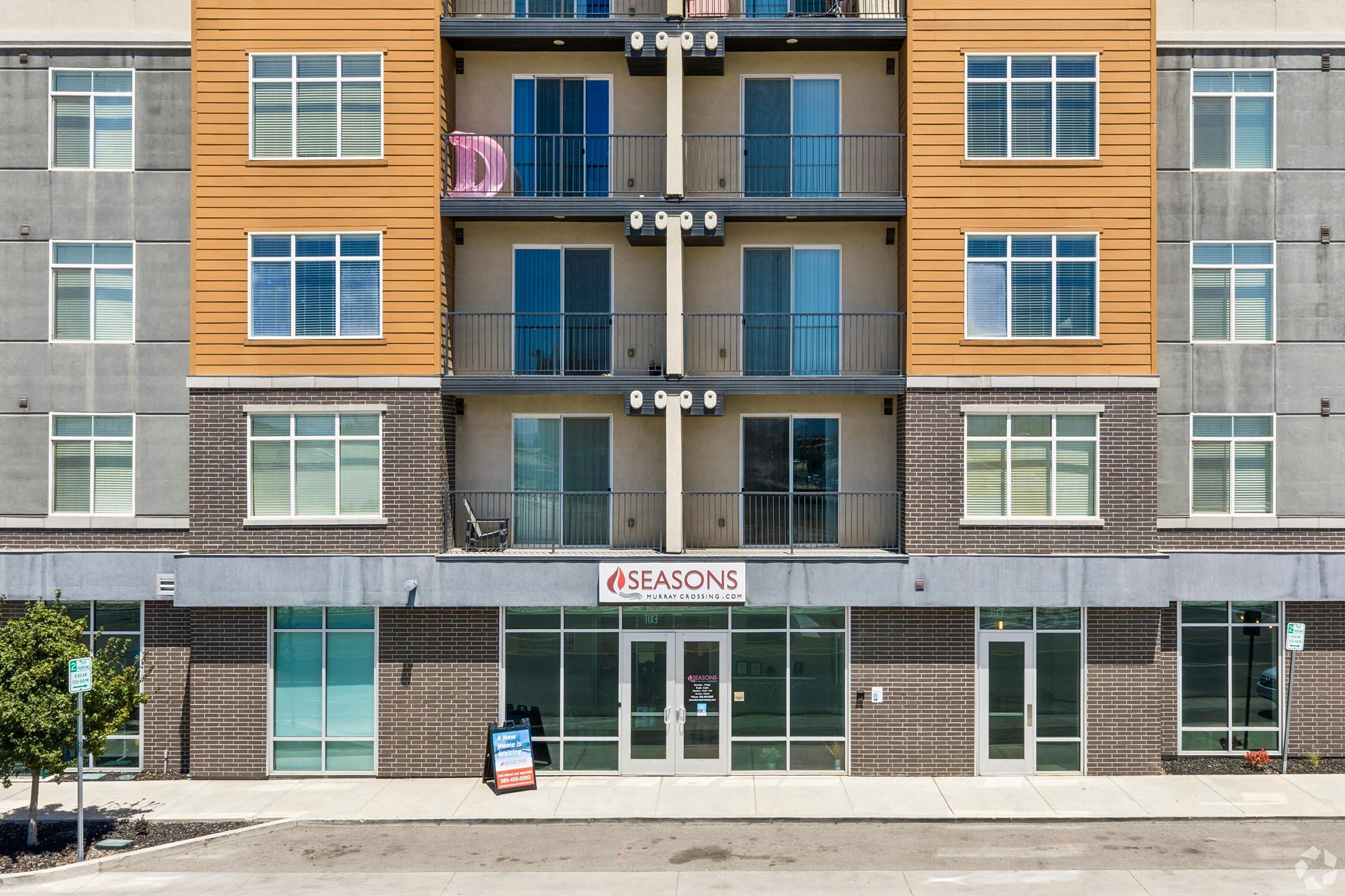 A modern apartment building featuring large windows and a brick facade. The lower level displays a sign for "SEASONS," and there are potted plants near the entrance. Balconies with railings are visible on the upper floors, some with furniture. The surroundings include parking and a landscaped area.