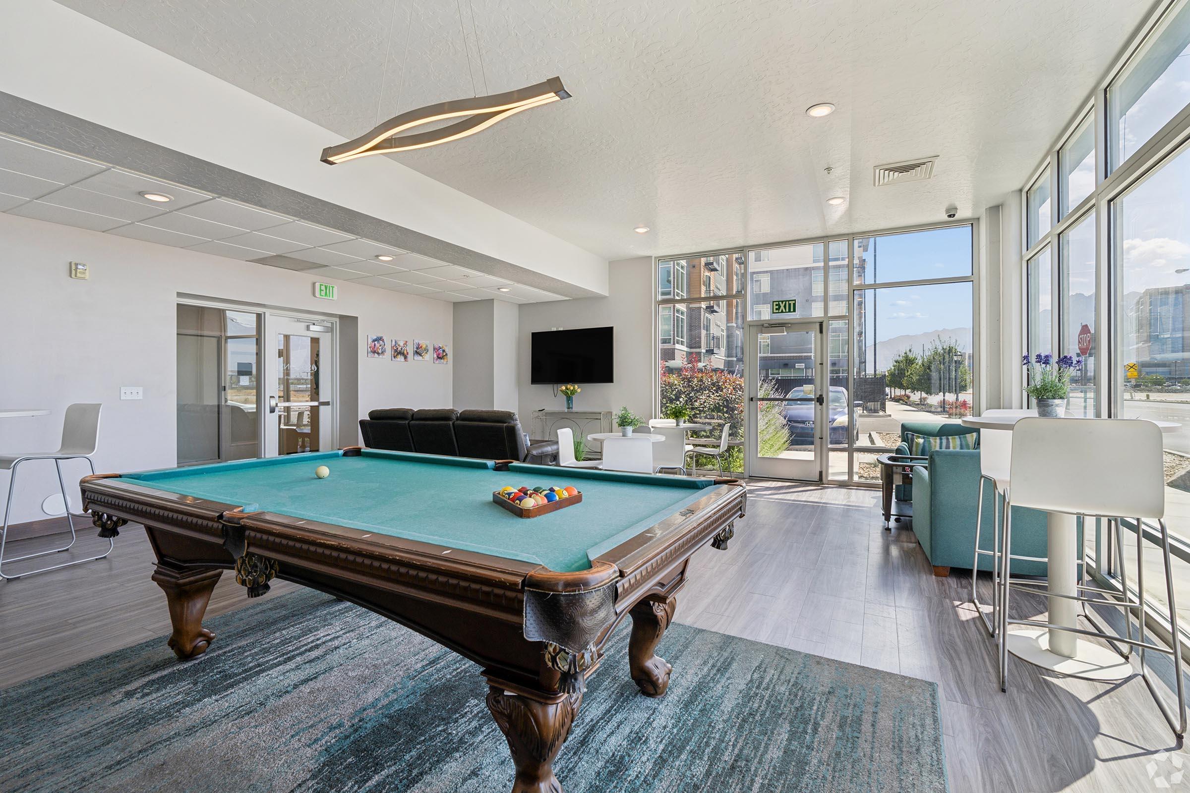 A modern lounge area featuring a billiard table with a green felt surface, surrounded by stylish seating and large windows allowing natural light. The space includes a television, decorative elements, and a view of an outdoor area, creating an inviting atmosphere for relaxation and socializing.