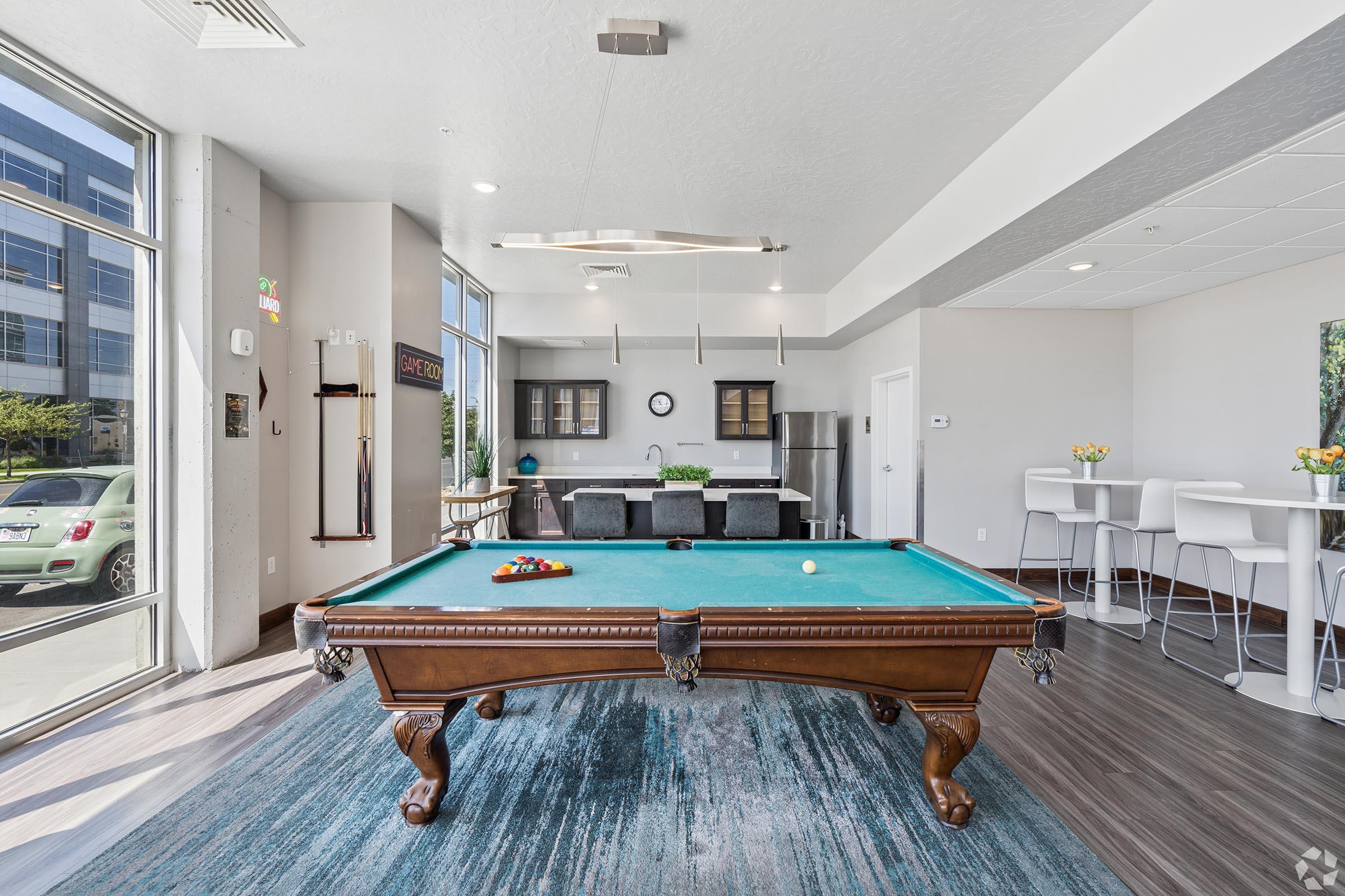 A modern pool room featuring a classic billiards table with a green felt surface. Natural light enters through large windows, illuminating the space. In the background, there's a kitchenette with dark cabinets, a dining area with stools, and decorative plants, creating a stylish and inviting atmosphere.