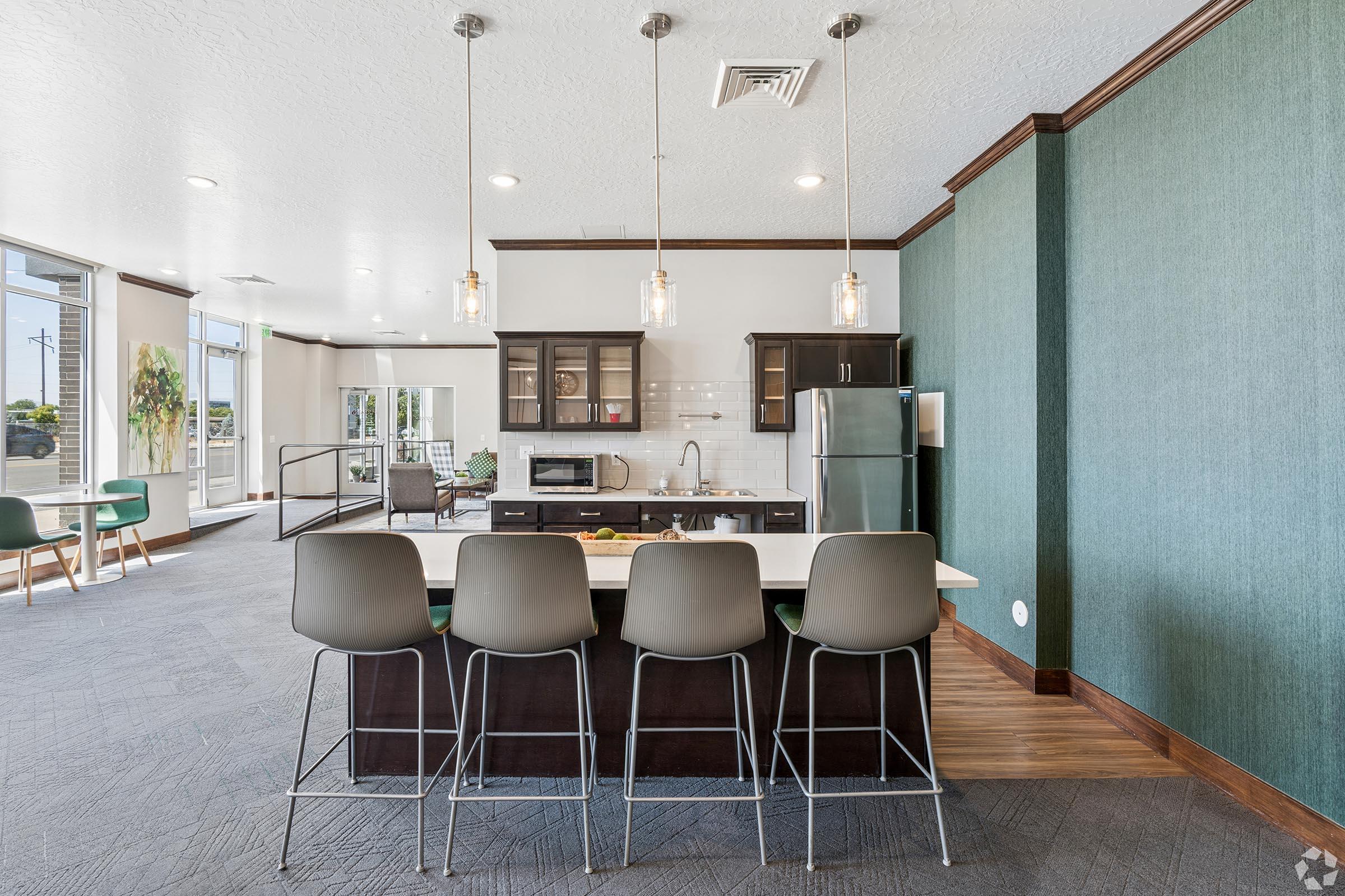 A modern kitchen area features a sleek island with four stylish bar stools. Behind the island, there are dark cabinets, a stainless steel refrigerator, and a microwave. The walls are a calming teal color, and large windows allow natural light to fill the space, creating a welcoming atmosphere.