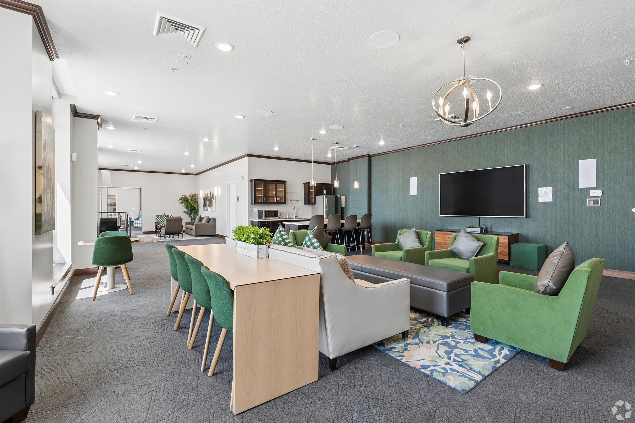 A modern lounge area featuring a variety of seating options, including green armchairs and a gray sofa. There is a communal dining table, a large television mounted on the wall, and a kitchenette in the background. Soft lighting and decorative plants add a welcoming atmosphere to the space.