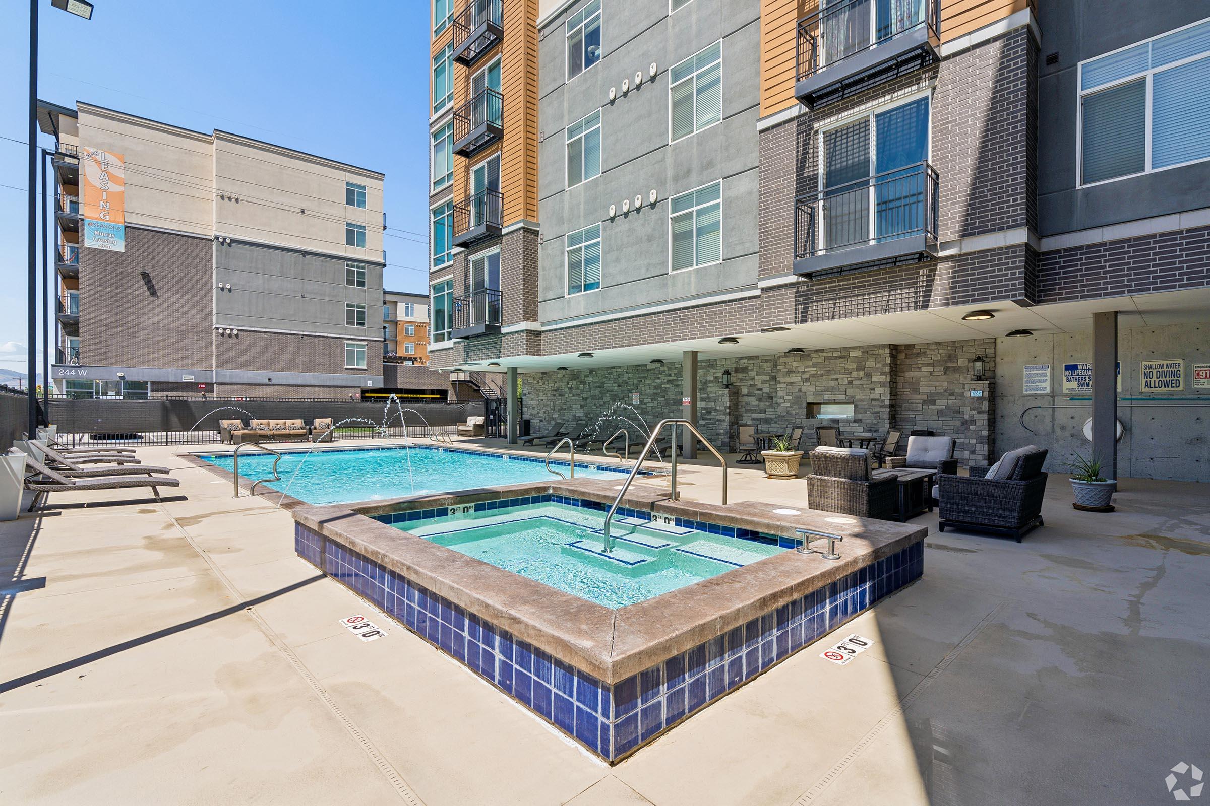 Outdoor swimming pool area with a large hot tub, surrounded by lounge chairs and modern apartment buildings. The pool features a clean, inviting design with clear blue water, and there are sun umbrellas providing shade. The area is well-lit and appears to be well-maintained, perfect for relaxation.