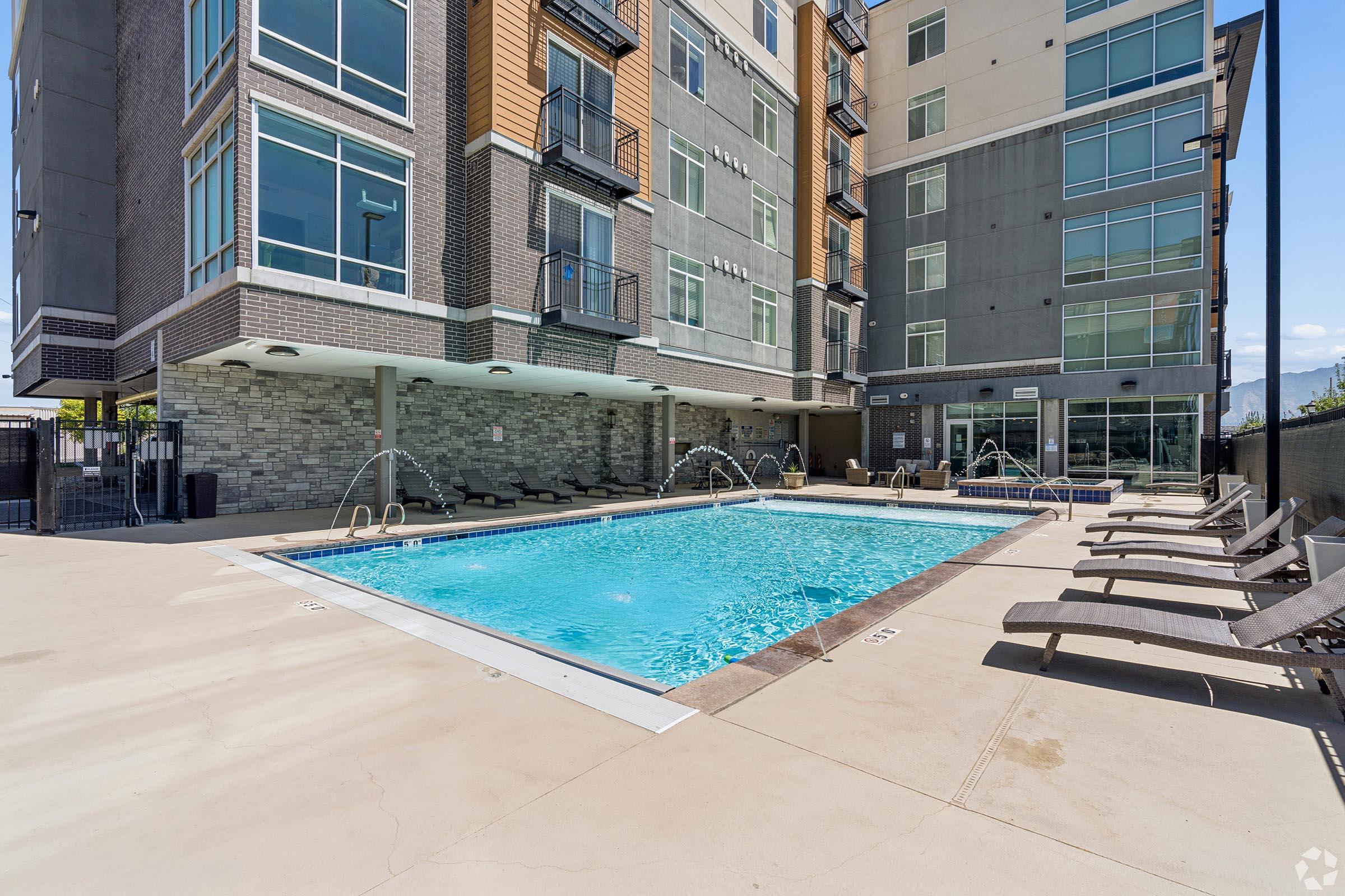 A modern apartment building with a swimming pool in the foreground. The pool is surrounded by lounge chairs and features decorative water fountains. The building has multiple stories with large windows, showcasing contemporary architectural design against a clear blue sky.
