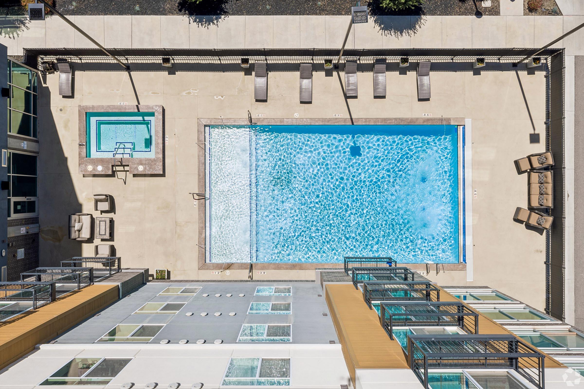 Aerial view of a swimming pool area featuring a large pool with clear blue water, surrounded by lounge chairs. There is also a smaller hot tub adjacent to the pool. The surrounding area includes elements of a modern building with balconies and a landscaped border.