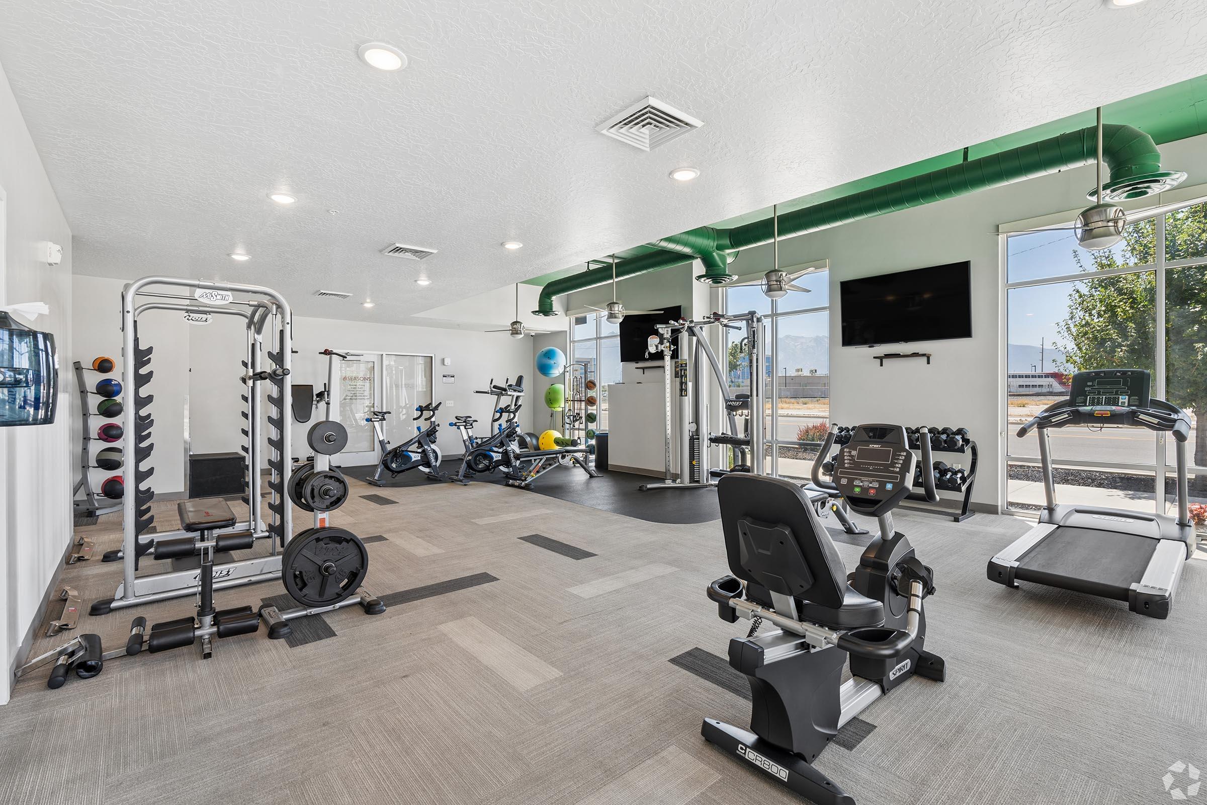 A modern gym interior featuring various exercise equipment, including a treadmill, stationary bikes, weight machines, and free weights. The space is well-lit with large windows and has a clean, organized layout. Green accents on the ceiling add a vibrant touch.