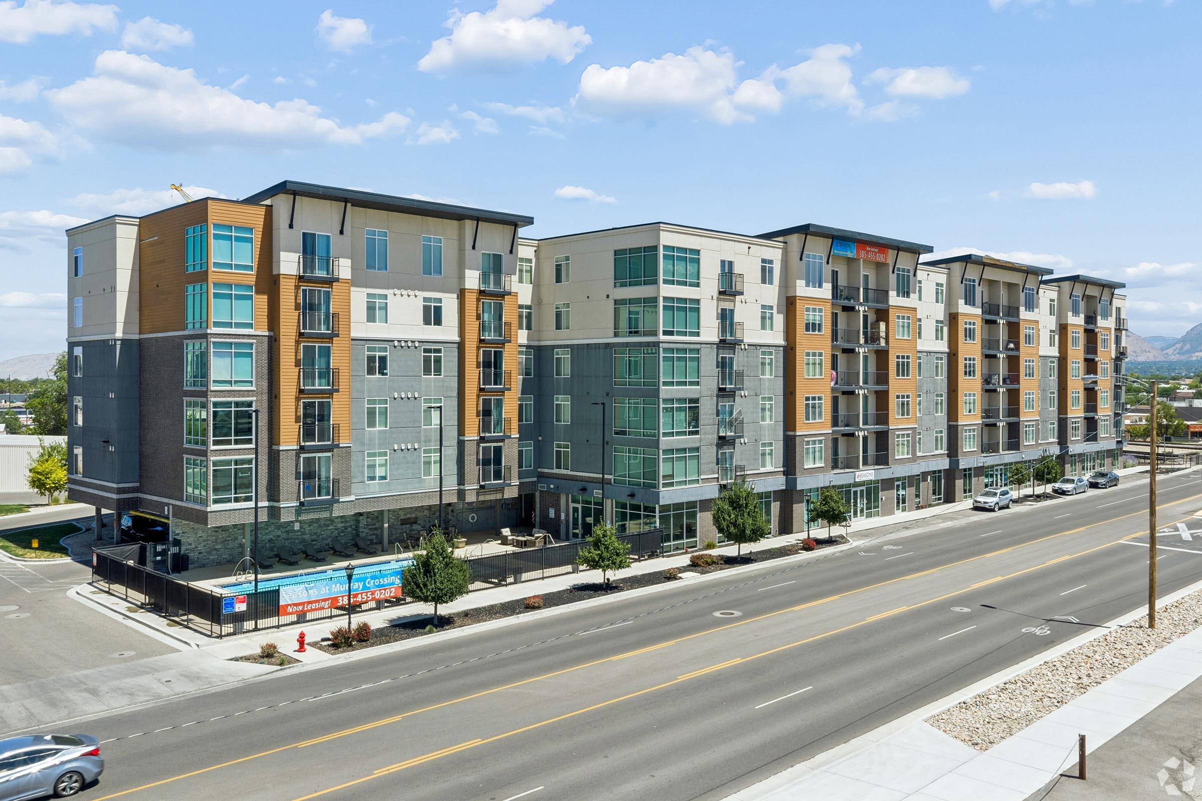 Modern apartment building with multiple stories, featuring a mix of brick and siding exteriors. The structure has balconies on upper floors, large windows, and is situated along a street lined with trees. A clear blue sky and scattered clouds are visible in the background, enhancing the urban landscape.