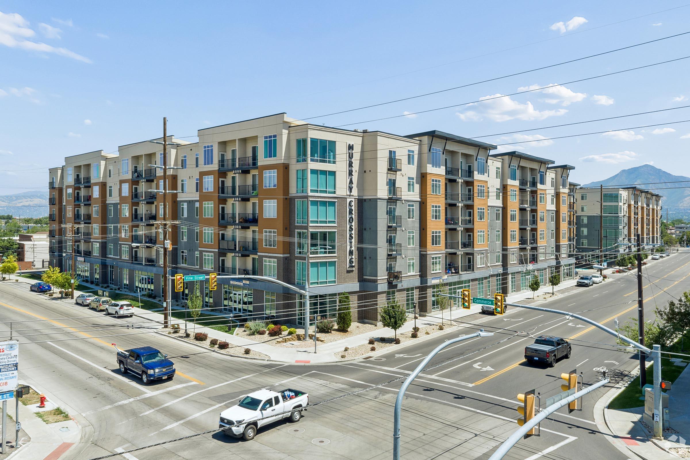 A modern multi-story apartment building situated at a busy intersection. The building features a mix of colorful exterior materials and has balconies on several floors. Surrounding streets are lined with vehicles, traffic signals, and greenery, with mountains visible in the background under a clear blue sky.