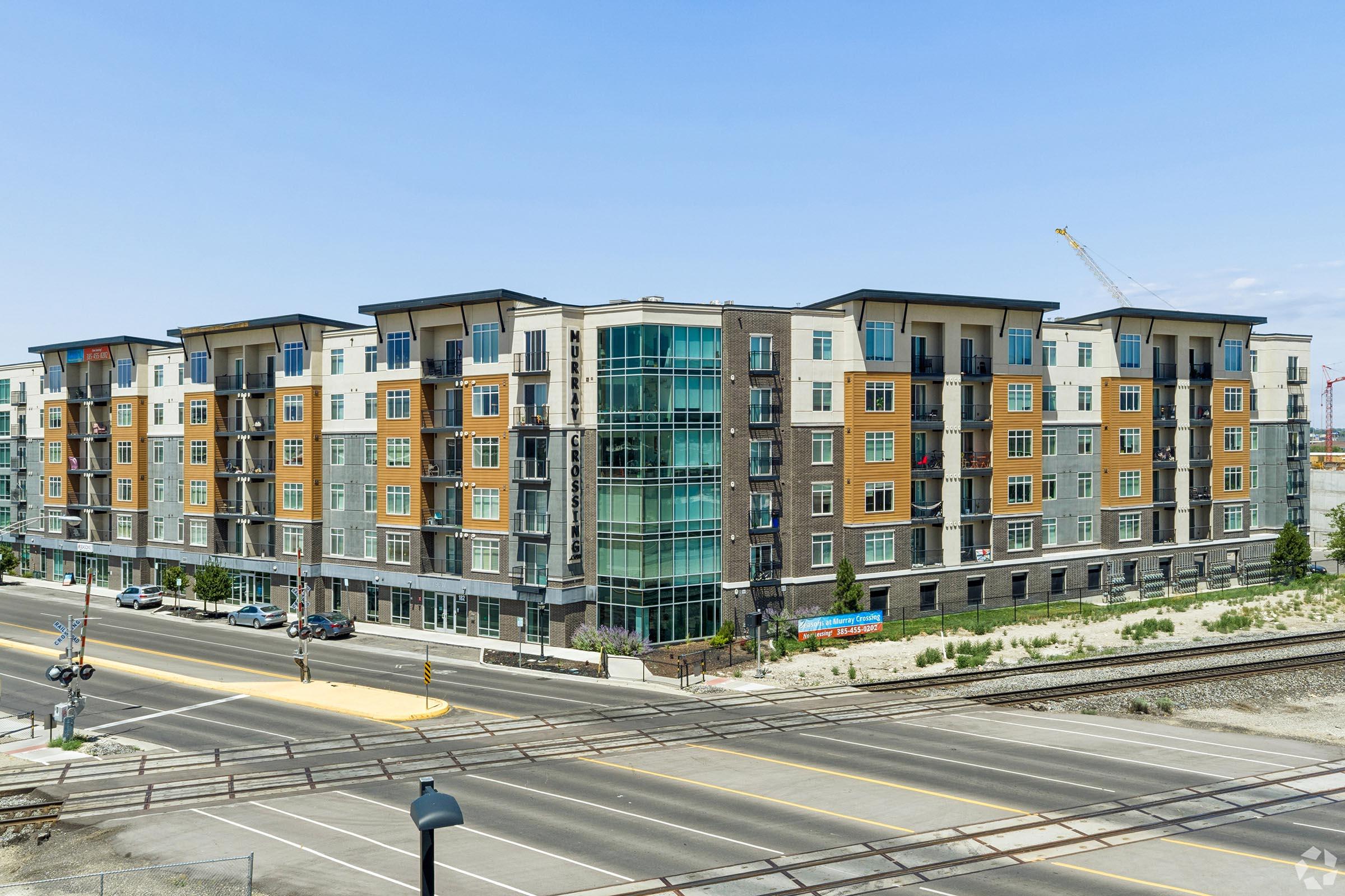 A modern multi-story apartment building with a mix of brick and colorful façade, situated near a street and railway tracks. The structure features numerous windows and balconies, with cars parked along the road. Cranes in the background suggest ongoing development in the area.