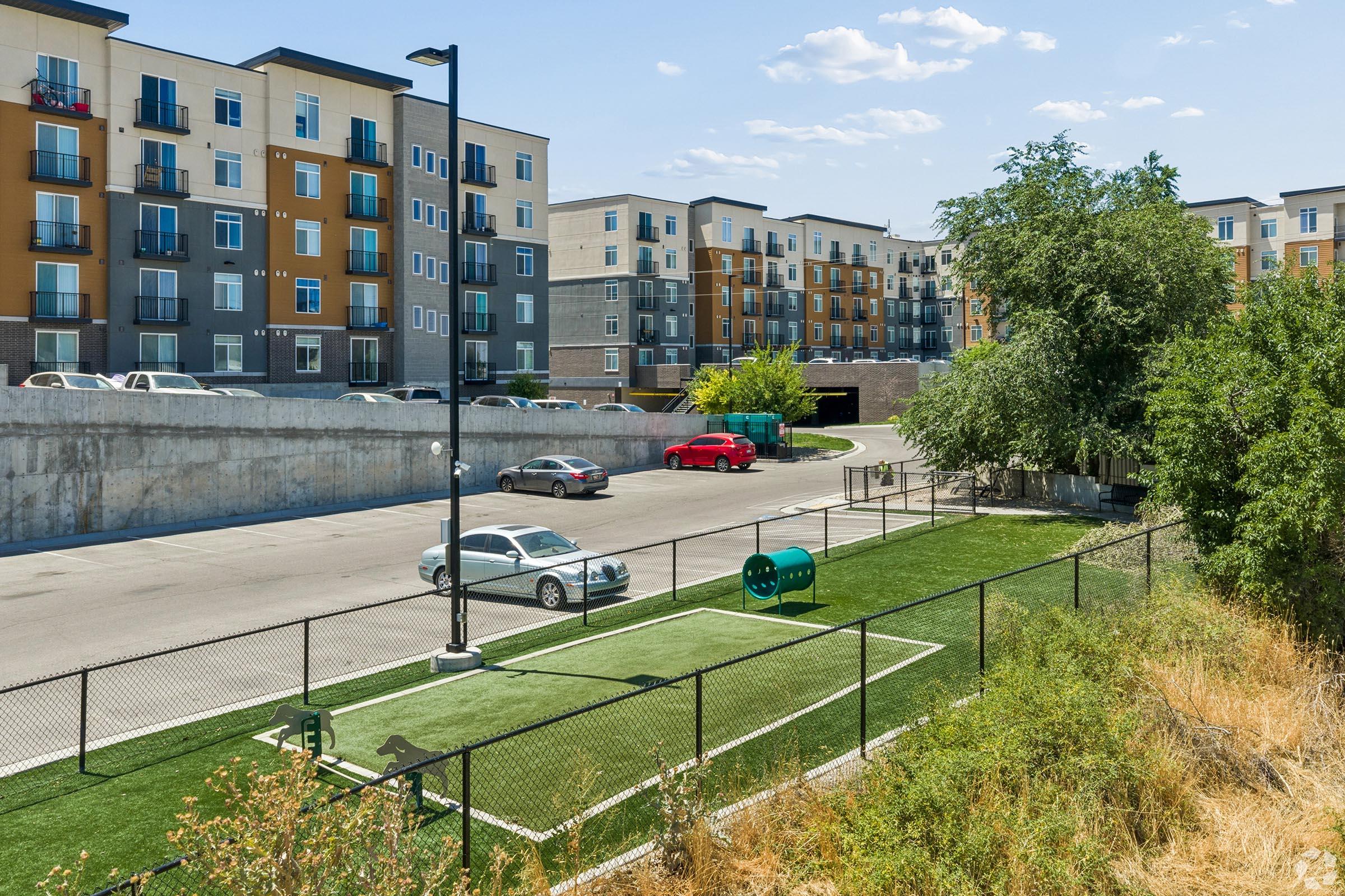 A green space with a small dog park featuring artificial turf and a dog waste station. Nearby, there are cars parked along a concrete area. In the background, modern apartment buildings with balconies are visible under a clear blue sky.