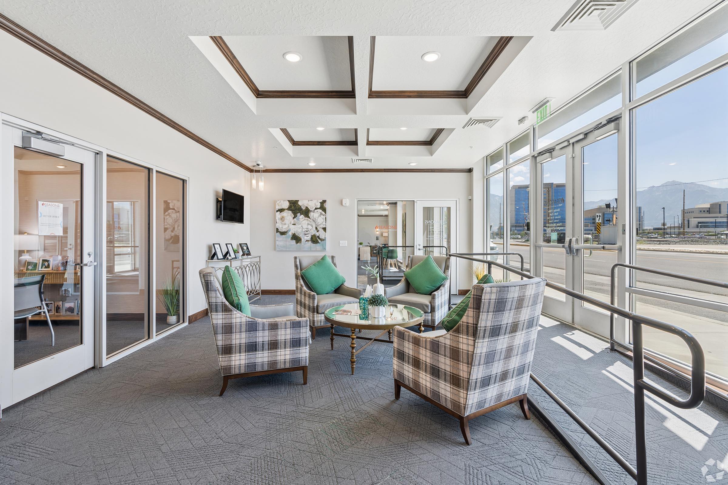 A bright, modern lounge area with two plaid armchairs adorned with green cushions, a small glass coffee table, and framed pictures on the walls. Large windows allow natural light to fill the space, showcasing a view of the outdoors. A door leads to another room, enhancing the open feel of the area.