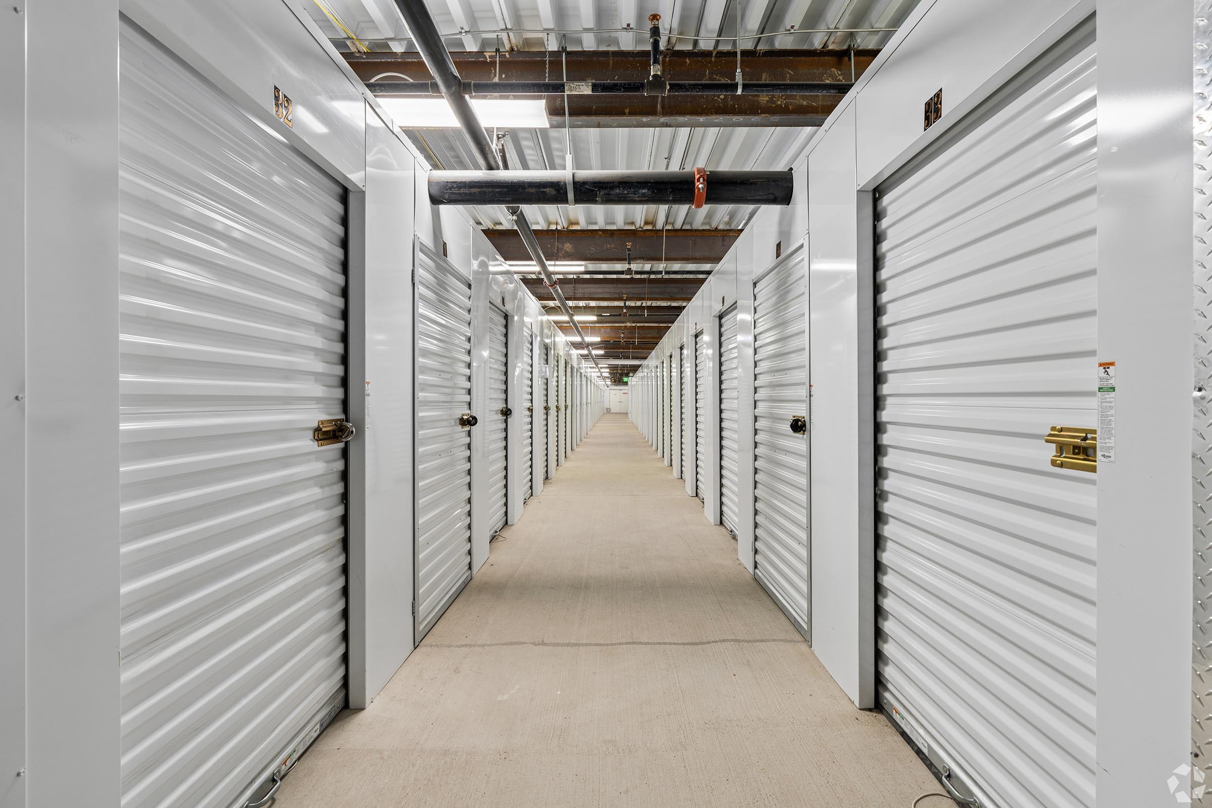 A narrow corridor lined with white storage units. Each unit has a metal roll-up door and a padlock. The ceiling features exposed pipes and metal beams. Bright lighting illuminates the space, creating a clean and organized appearance. The floor is concrete, and the corridor extends further into the background.