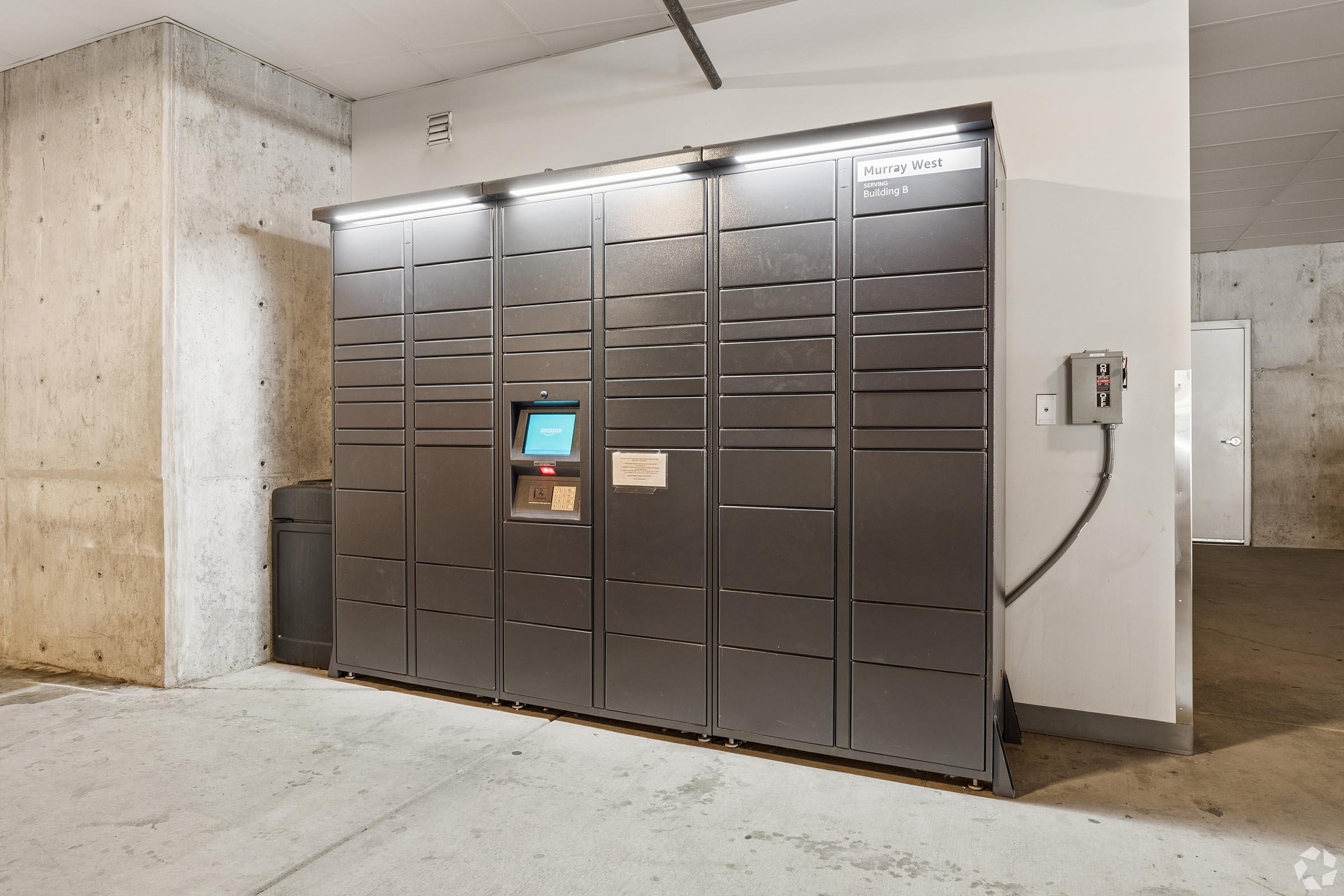 A large, gray package locker system located in a concrete area. The locker unit features a digital screen for user interactions. Nearby, there is a power outlet on the wall, and a trash can is visible in the background. The space appears clean and well-lit, indicative of an indoor building environment.