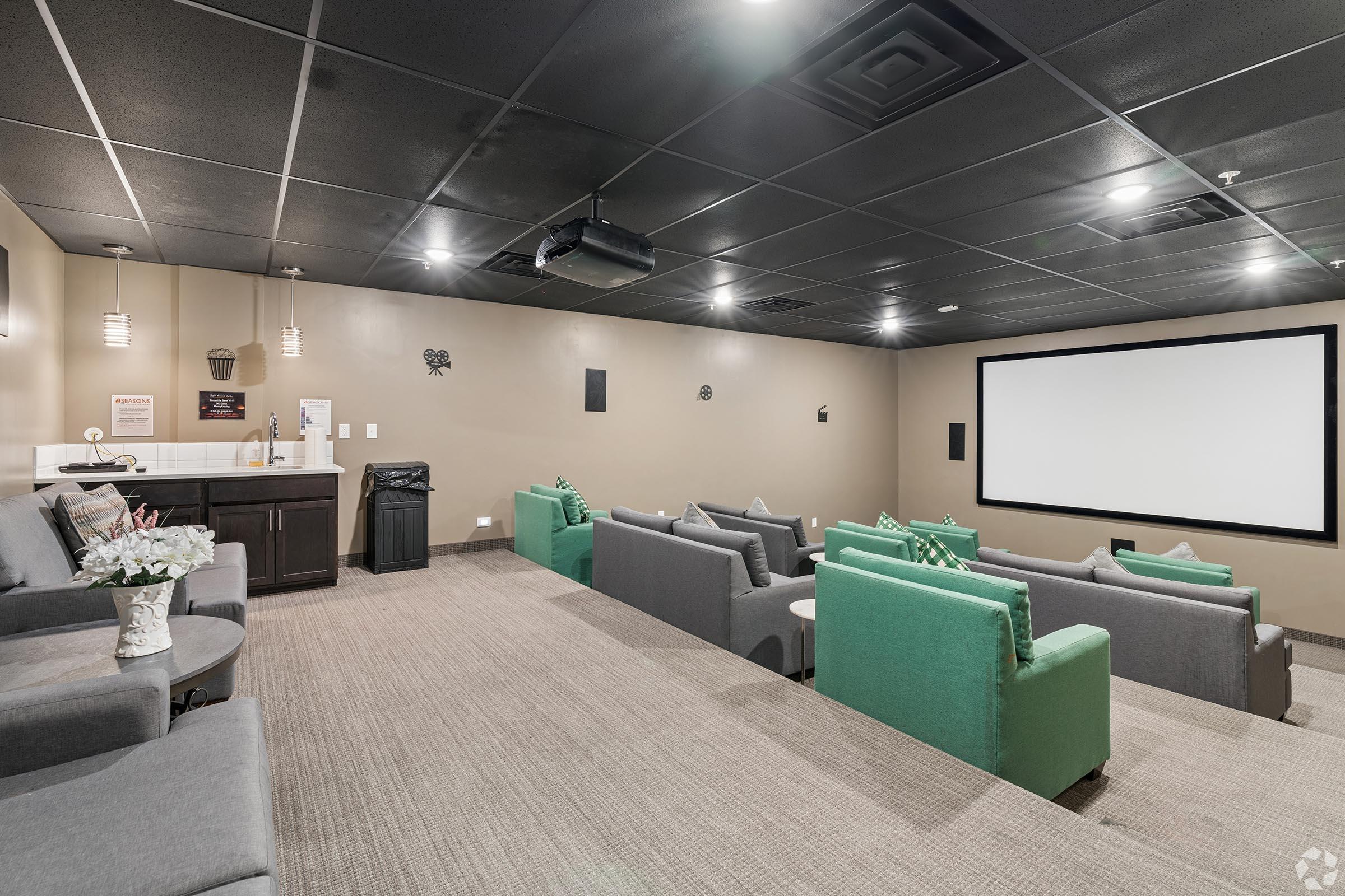 A cozy home theater featuring comfortable gray couches with green accents, a large projector screen at the front, and a small snack area on the side. The room has a modern design with dim lighting, perfect for movie nights or presentations.