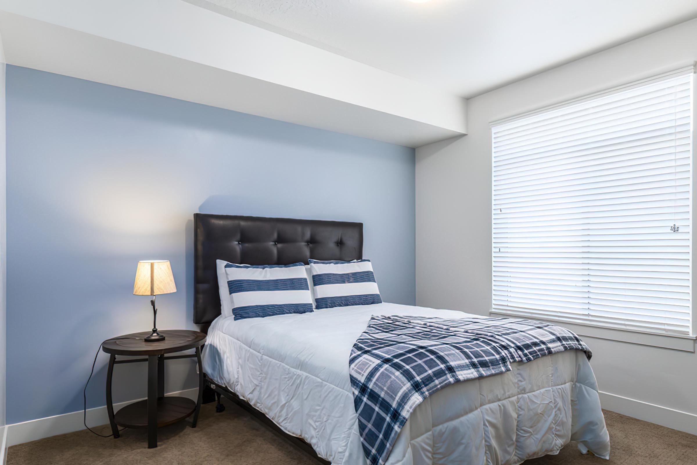A cozy bedroom featuring a dark upholstered headboard with a white and blue plaid comforter on the bed. There’s a bedside table with a lamp, and the room has light blue walls and a window with white blinds, allowing natural light to fill the space.