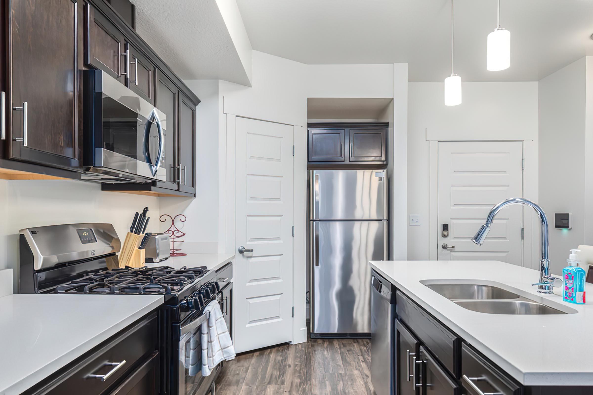 Modern kitchen featuring dark wood cabinetry, stainless steel appliances including a microwave, oven, and refrigerator. A gas stove is positioned on the left with utensils nearby, and a sink centered on the island. Bright overhead lights and a white countertop enhance the spacious, contemporary design.