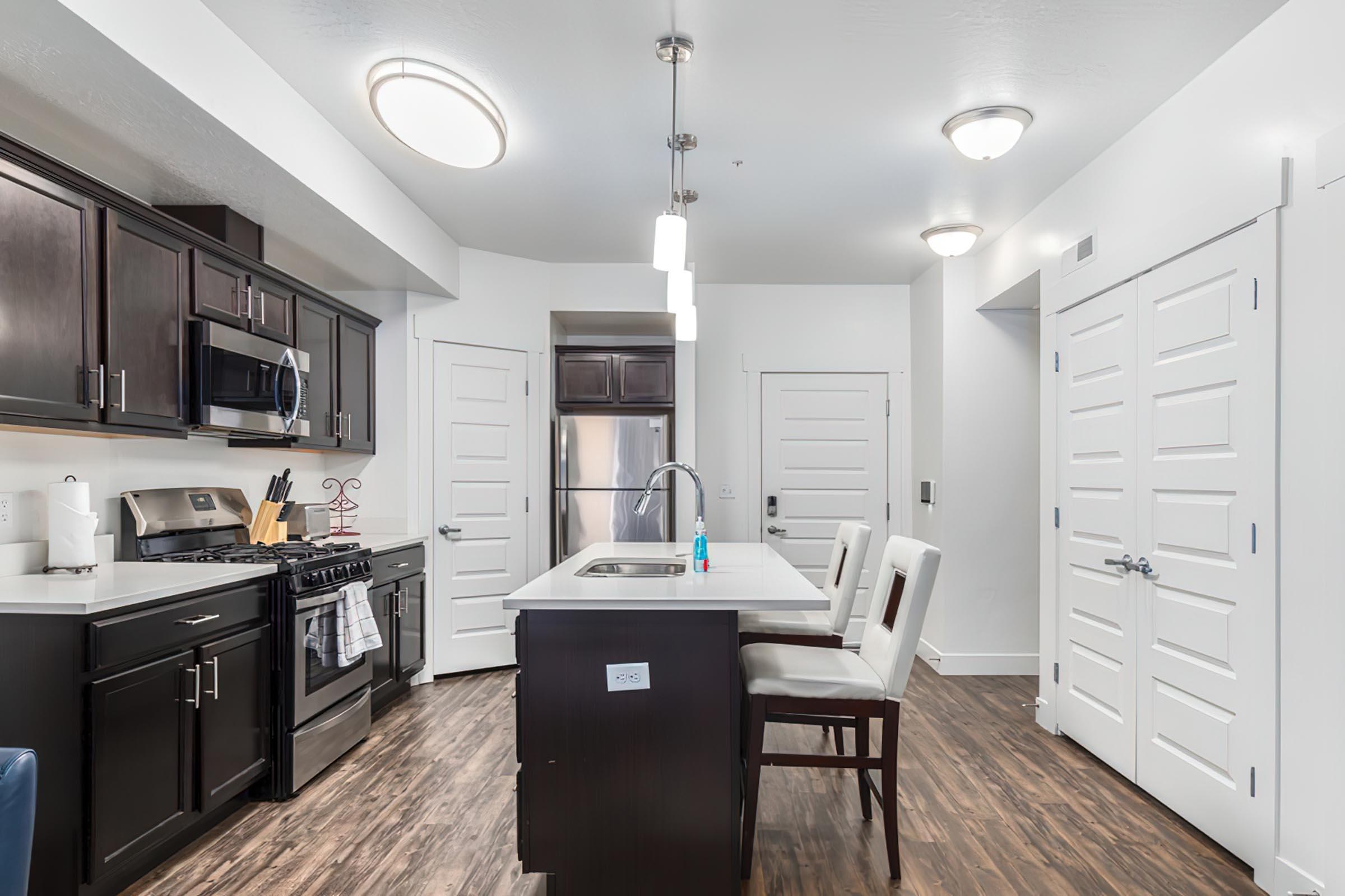 Modern kitchen featuring dark cabinetry, stainless steel appliances, and a central island with seating for two. The space is well-lit with ceiling lights and natural light from a nearby window. There are white doors leading to other areas of the home, and the flooring is wood-like laminate.
