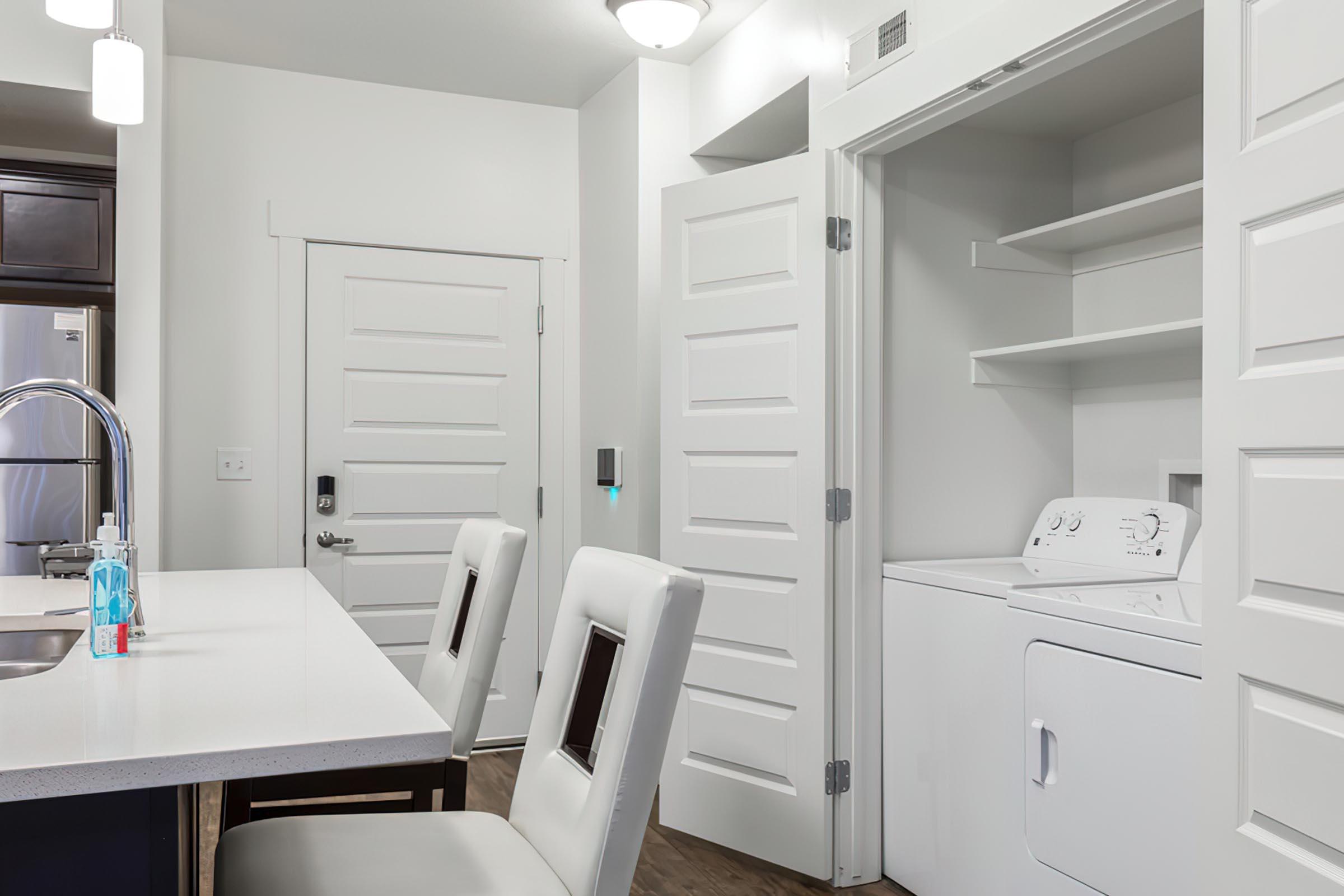 A modern kitchen with a white countertop and two padded bar stools. On the right, there is a laundry area with a washing machine tucked into a closet space. The walls are painted white, and there are several doors leading to other areas. The lighting is bright and the space has a clean, contemporary look.