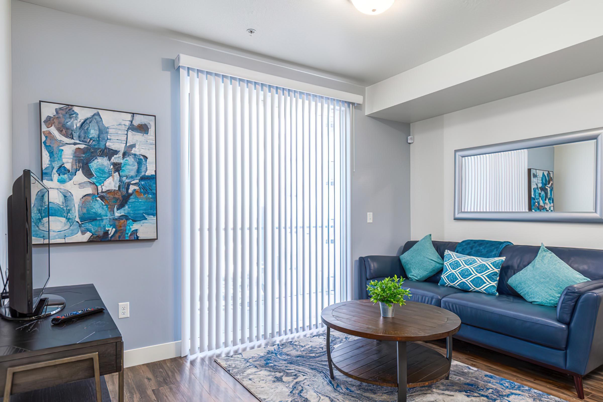 A modern living room featuring a blue couch with decorative cushions, a wooden round coffee table, and a TV on a stand. The walls are painted light gray, with a colorful artwork piece above the couch and a large mirror on the opposite wall. Vertical blinds cover a window, allowing natural light to enter.