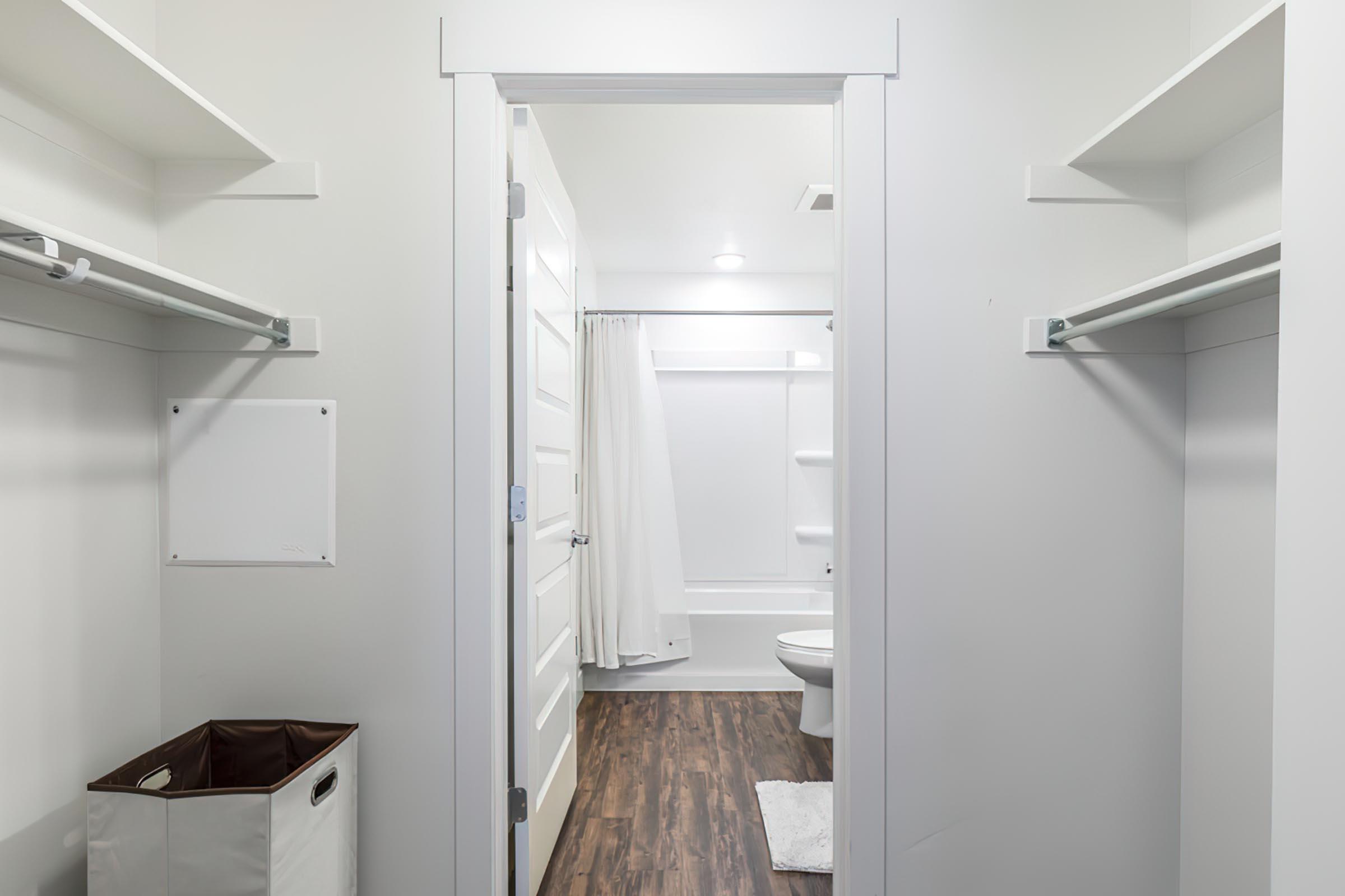 A view from a hallway into a clean, modern bathroom. To the left, there is a closet space with open shelves. The bathroom features a shower stall with a white curtain and a toilet, along with light-colored walls and a dark wood floor. A laundry basket is visible near the entrance.