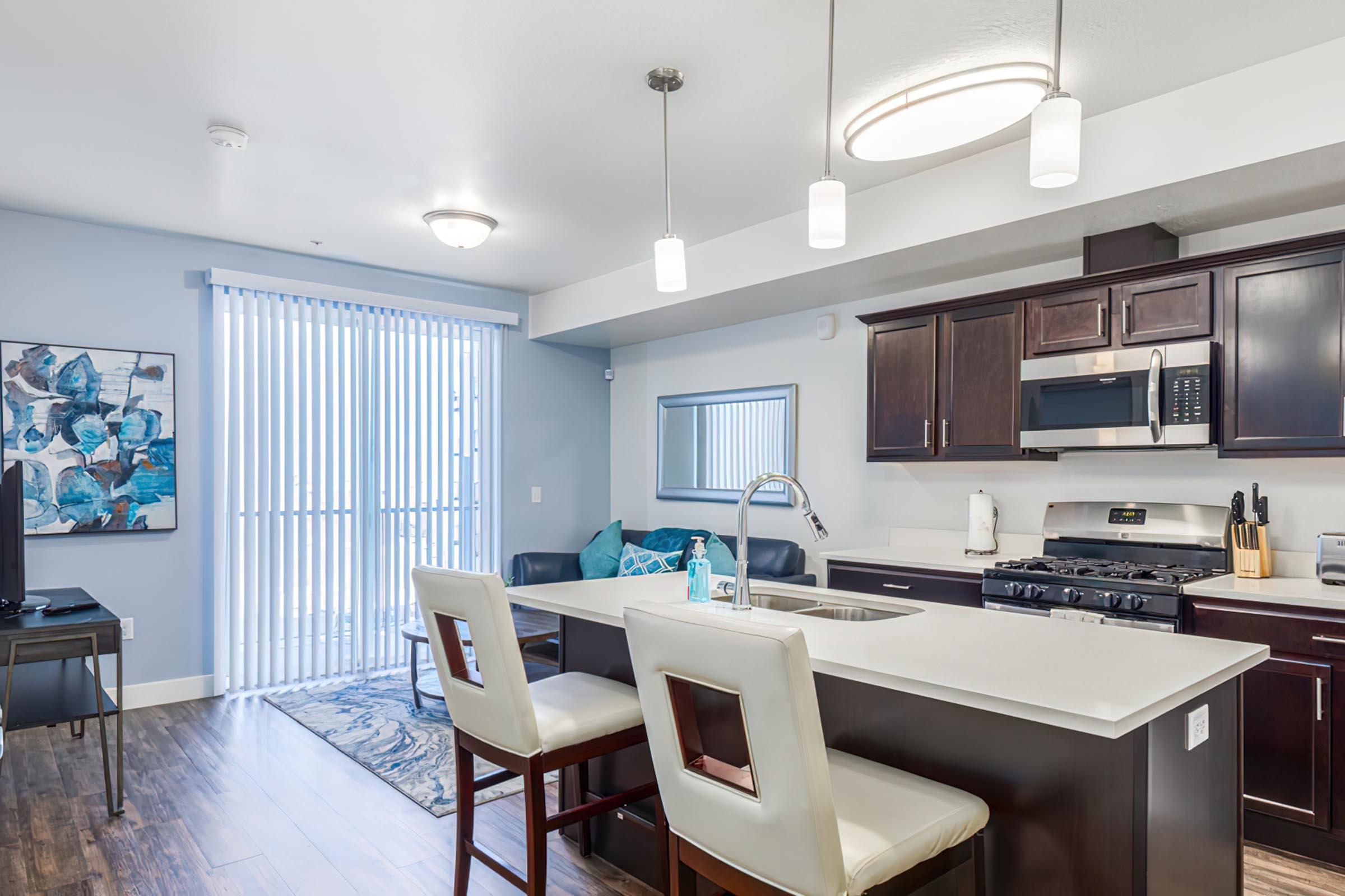 Modern kitchen and living area in an apartment with a large island, bar stools, dark wood cabinets, and stainless steel appliances. There is a dining area with a blue and white artwork on the wall, a television in the corner, and sliding glass doors leading to a balcony, illuminated by natural light.