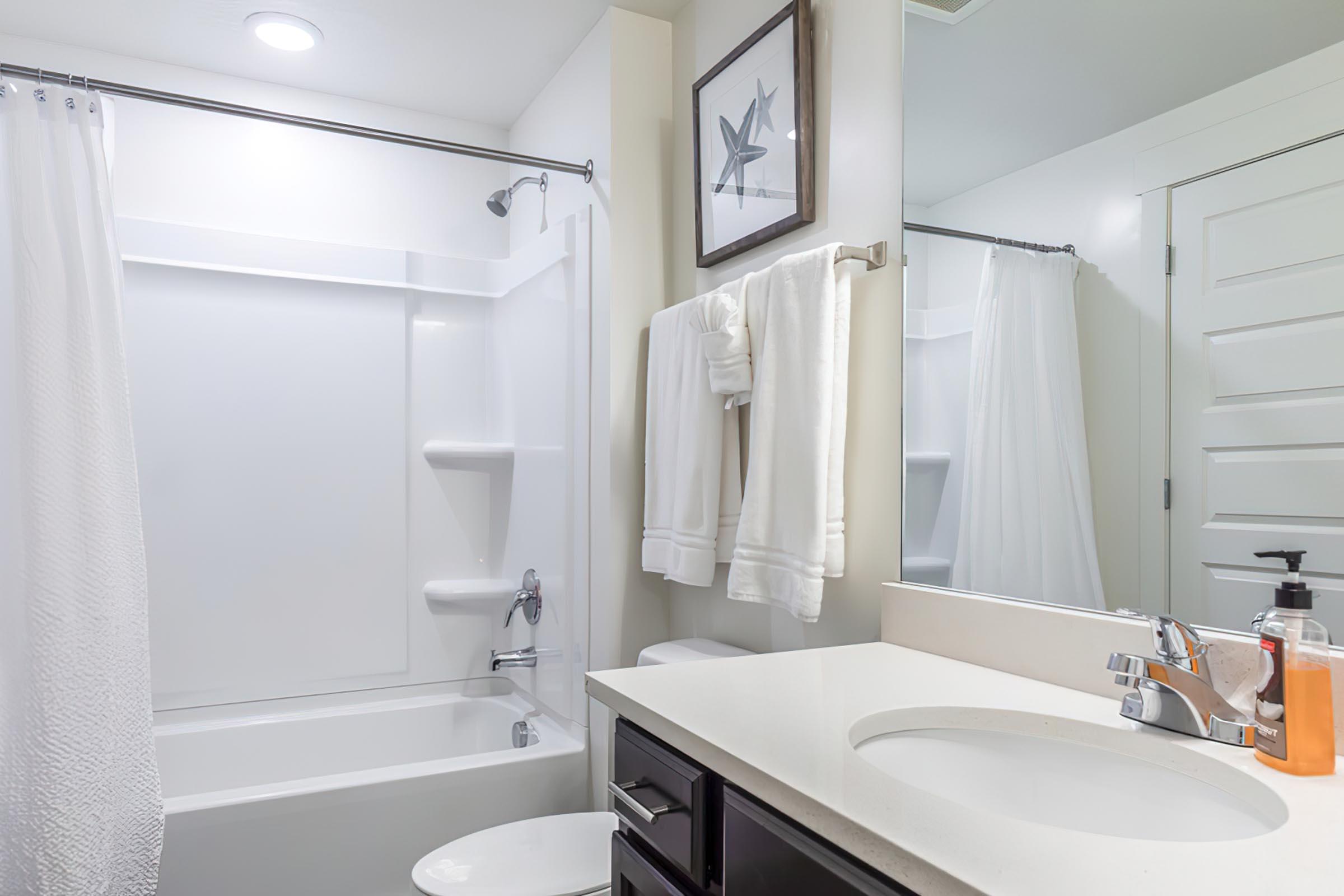 A modern bathroom featuring a bathtub with a shower, white curtains, a large mirror, a sink with a countertop, and dark cabinetry. There are neatly hung white towels and a soap dispenser on the counter, creating a clean and inviting atmosphere.