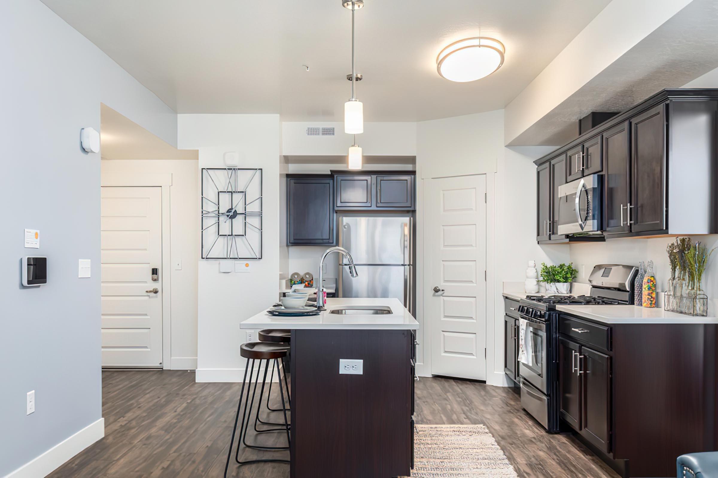 Modern kitchen featuring dark wood cabinetry, stainless steel appliances, and a central island with bar seating. The walls are painted light gray, and there is a decorative wall clock. A door leads to another room, and the floor has a dark wood finish, creating a contemporary and inviting atmosphere.