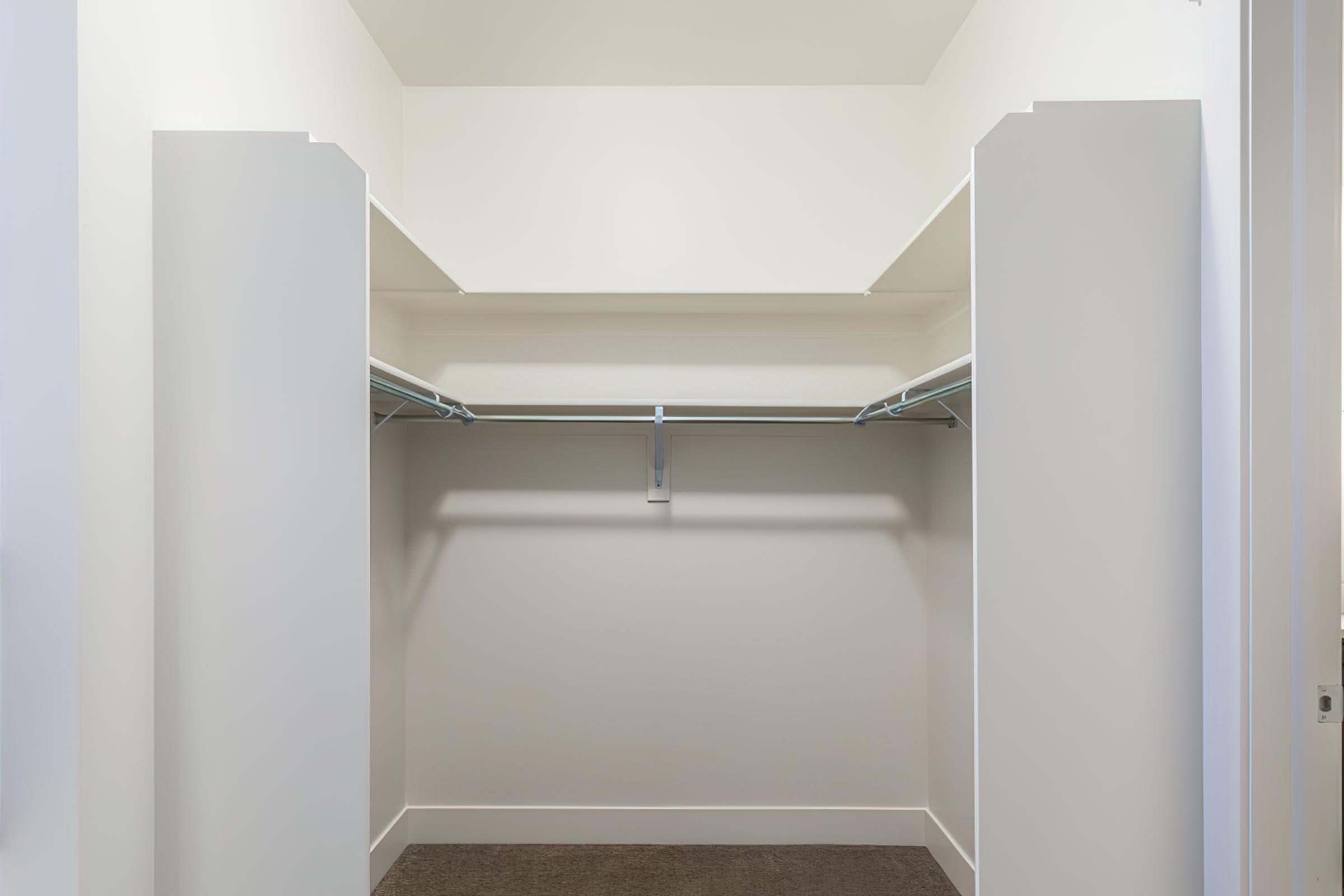 A spacious, empty walk-in closet featuring white walls and a light-colored carpet. The closet has two open shelving units with hanging rods installed, providing ample storage space, and is well-lit with natural light coming from an outside source.