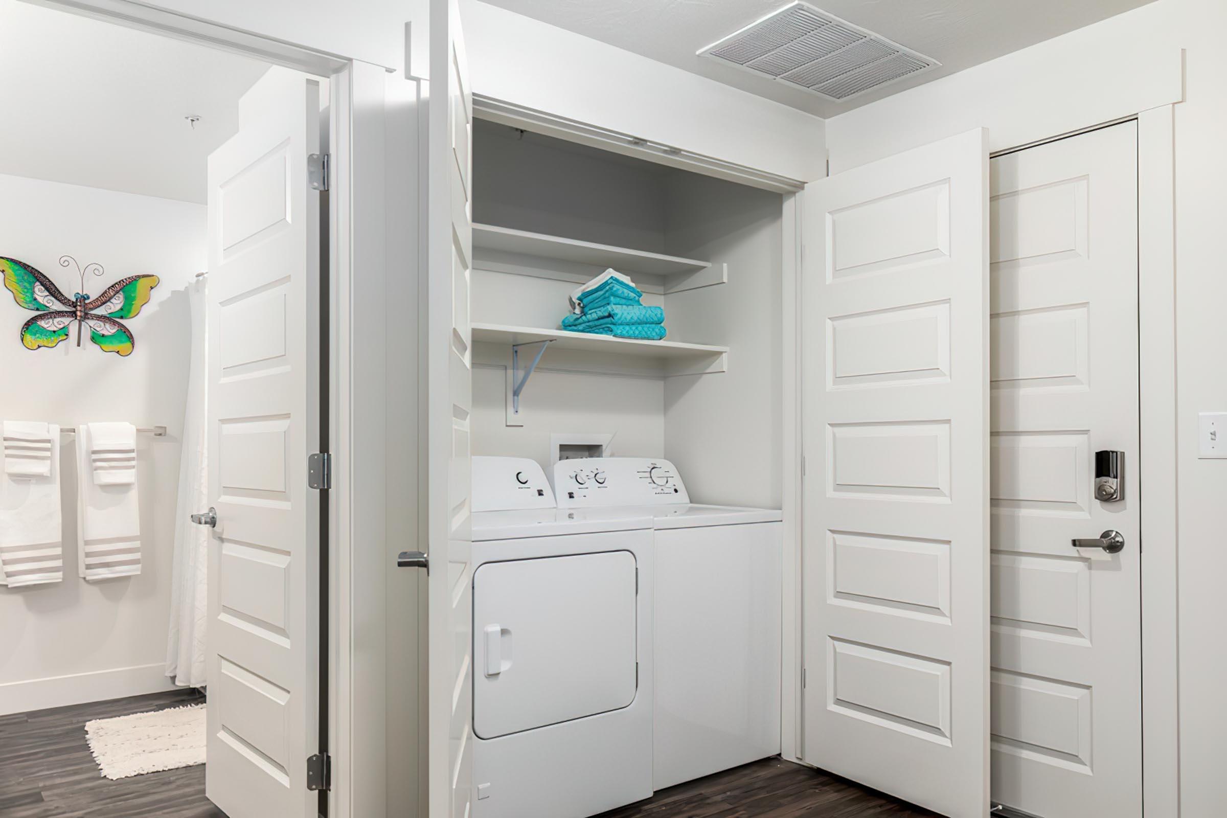 A laundry area featuring a stacked washer and dryer with open shelving above for storage. The room has light-colored walls and doors, and a butterfly wall decoration is visible. Towels hang on a hook nearby, and a door leads to another room.
