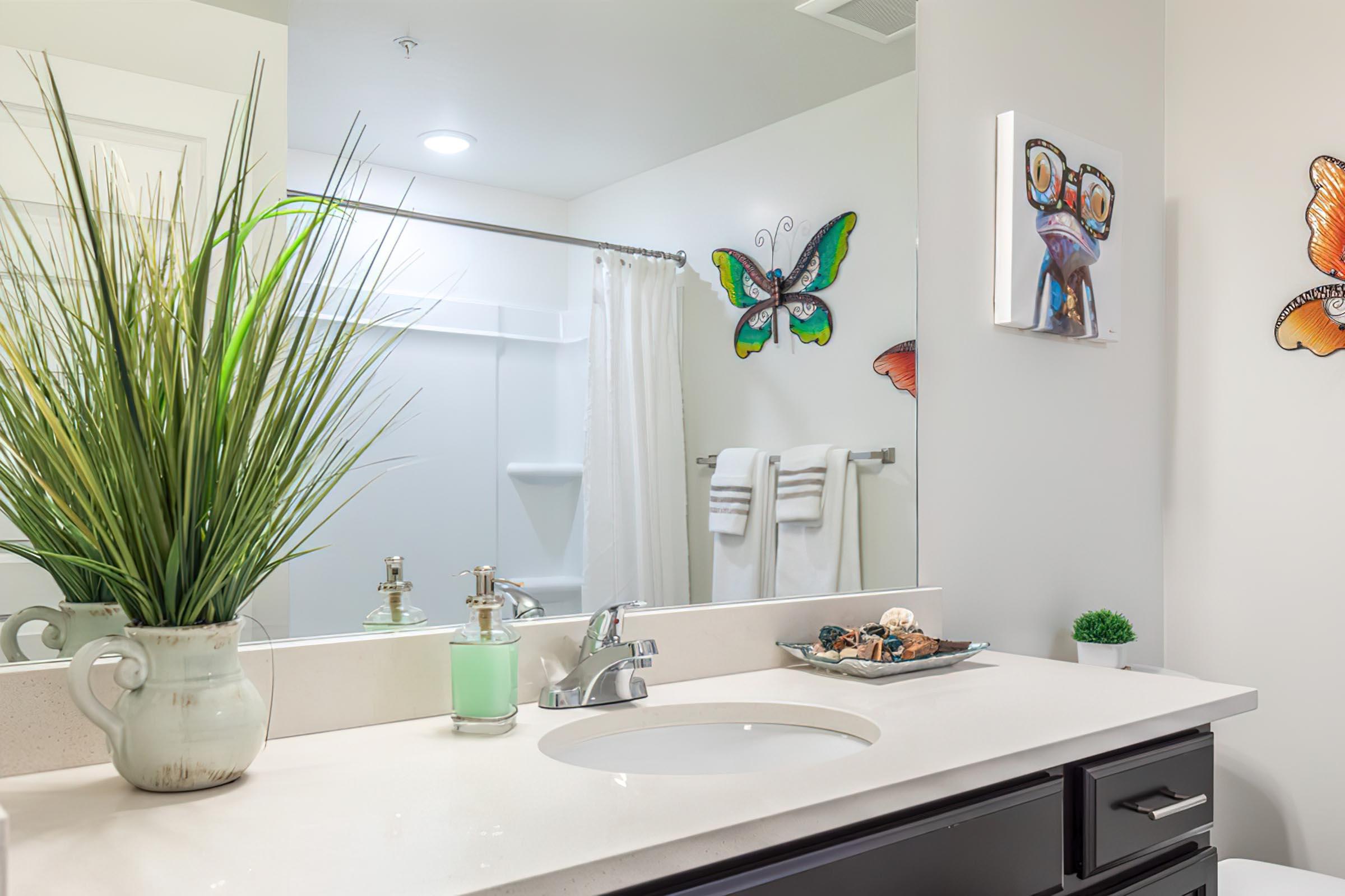 A bright and modern bathroom with a simple white countertop and sink. A large mirror reflects the space. Decorative elements include colorful butterfly wall art, green potted plants, and a small tray with decorative items. Soft white towels hang on a rack, adding to the inviting atmosphere.