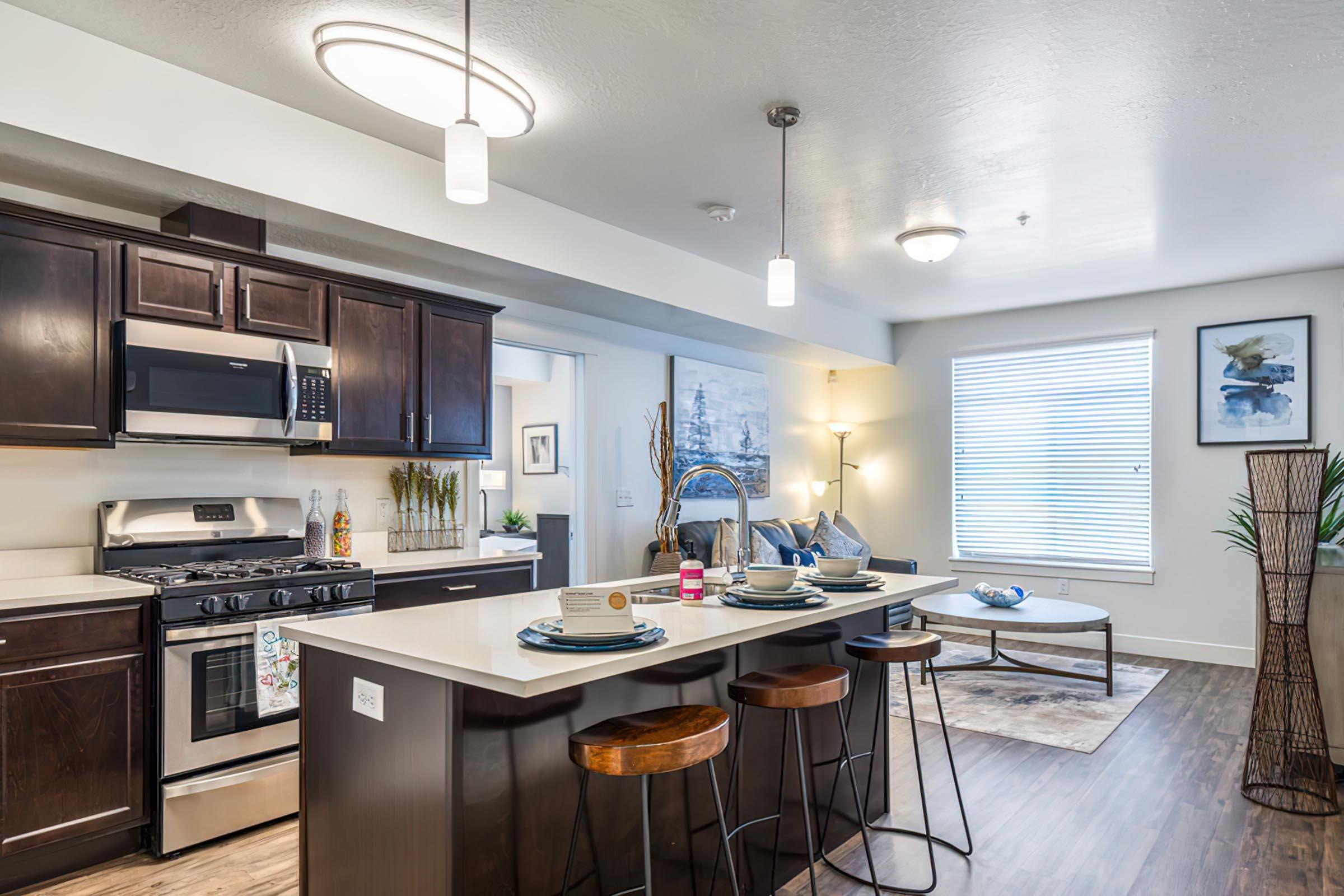 Modern kitchen and living area featuring dark wood cabinetry, stainless steel appliances, and a large island with seating. The space includes a dining setup, decorative plants, wall art, and natural light from a window, creating a welcoming and stylish ambiance.
