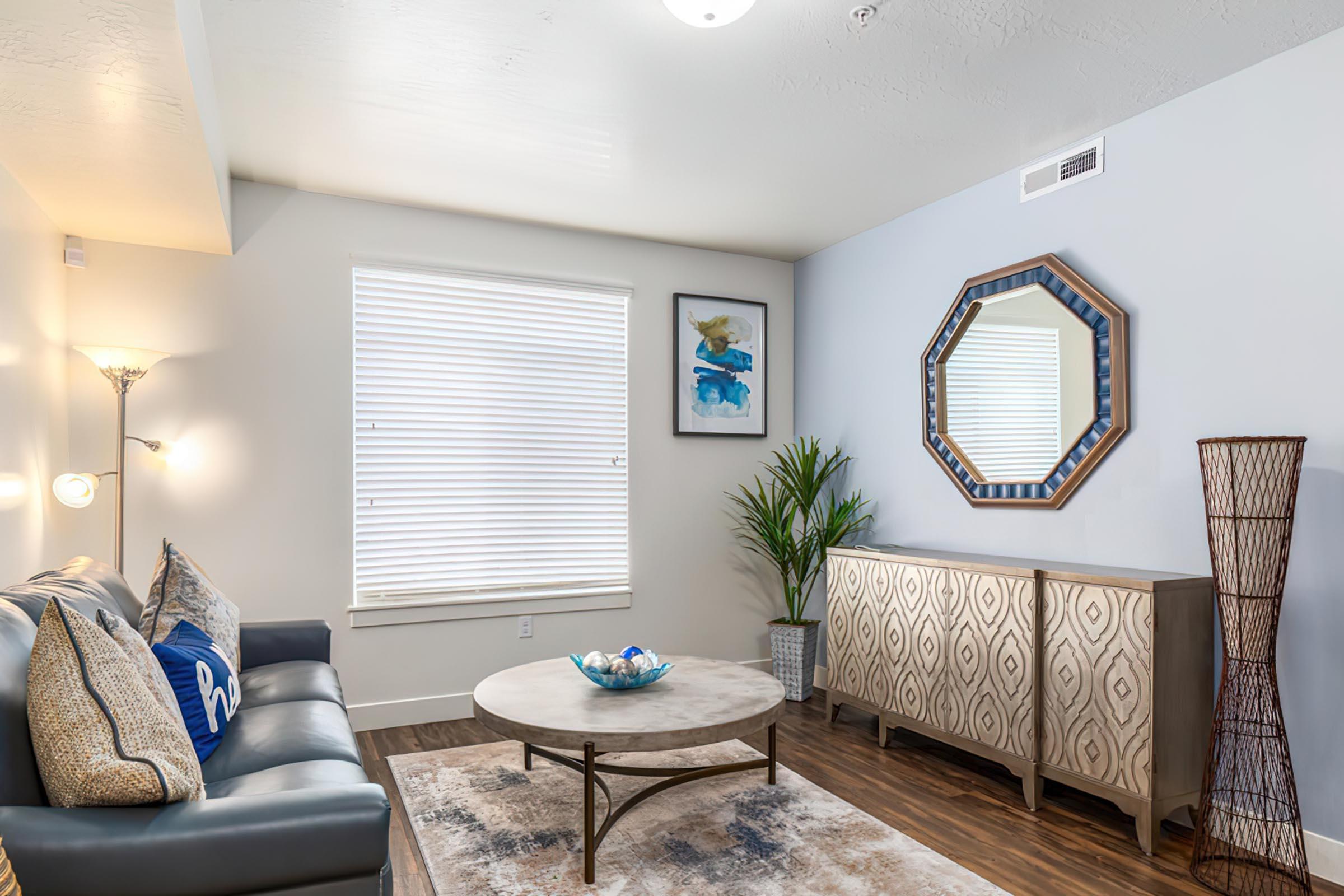A modern living room featuring a dark sofa with decorative pillows, a round coffee table, and a patterned cabinet. A tall plant adds greenery, and a framed artwork hangs on the wall. Natural light streams in through white blinds, illuminating the space. A decorative mirror complements the contemporary decor.
