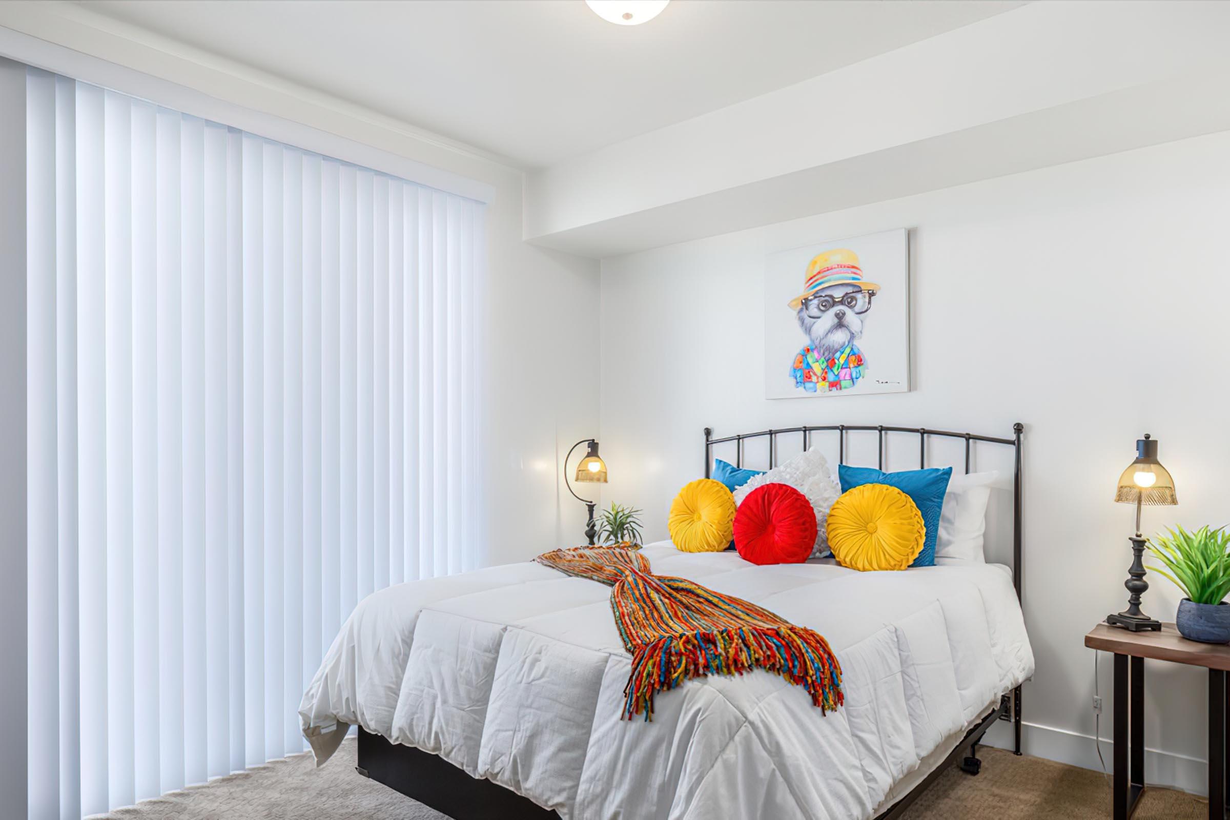 A modern bedroom featuring a black metal bed with a white comforter and colorful decorative pillows. A vibrant artwork of a dog wearing a hat hangs on the wall. The room has soft lighting from two bedside lamps, a small plant on a nightstand, and vertical blinds allowing natural light.