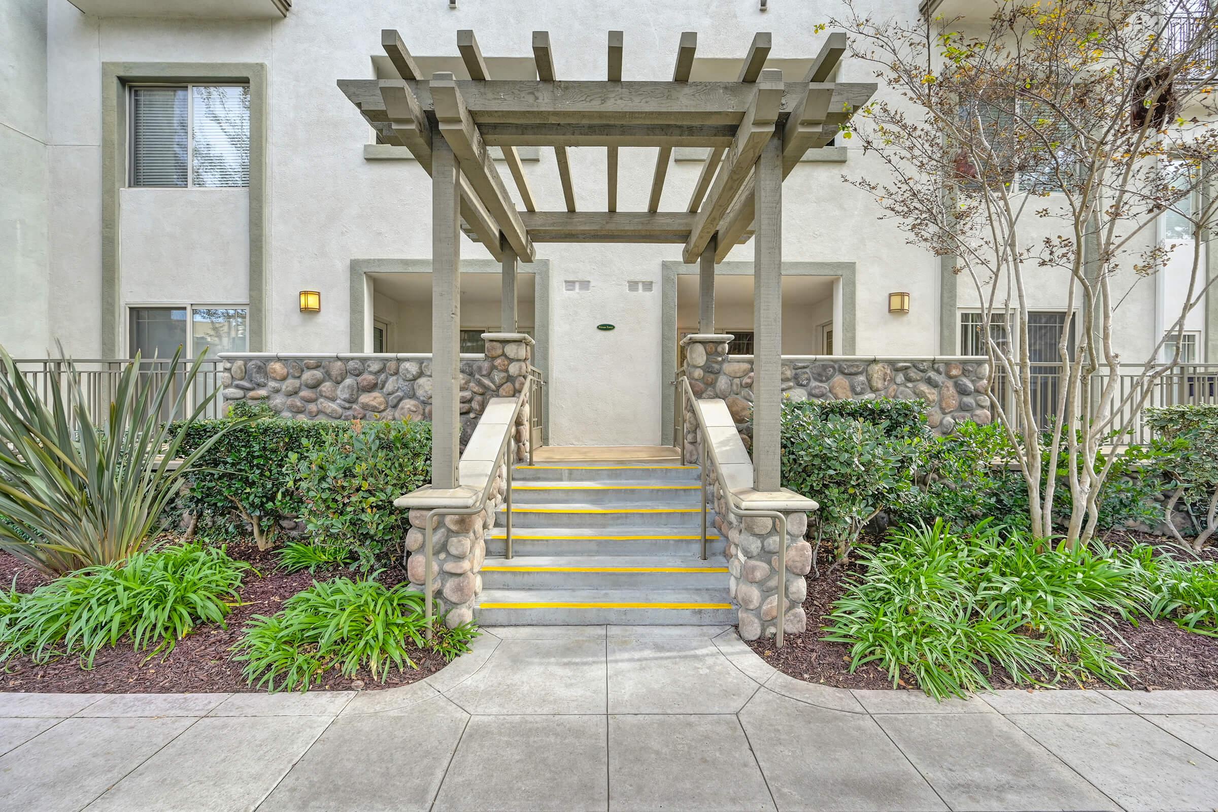 A well-maintained entrance with a decorative pergola and stone accents. The walkway features tiered stairs leading to a door, surrounded by lush green plants and landscaping. The building exterior is light-colored, with large windows and a welcoming atmosphere.