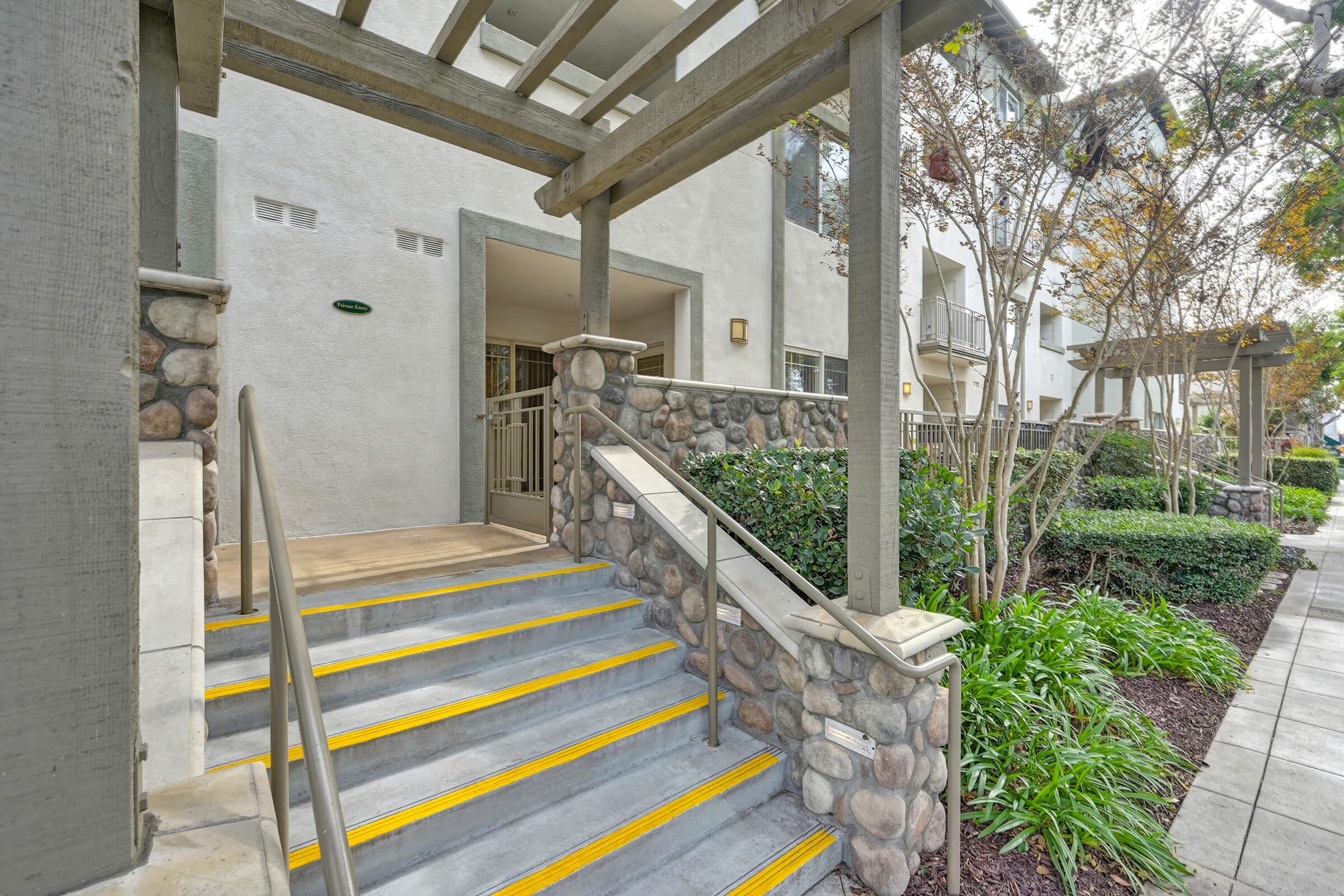 Exterior view of a building entrance featuring a set of steps with yellow markings, surrounded by landscaped greenery. The entrance is framed by wooden beams and stone walls, providing a welcoming atmosphere. Nearby foliage includes bushes and small trees, contributing to a well-maintained environment.