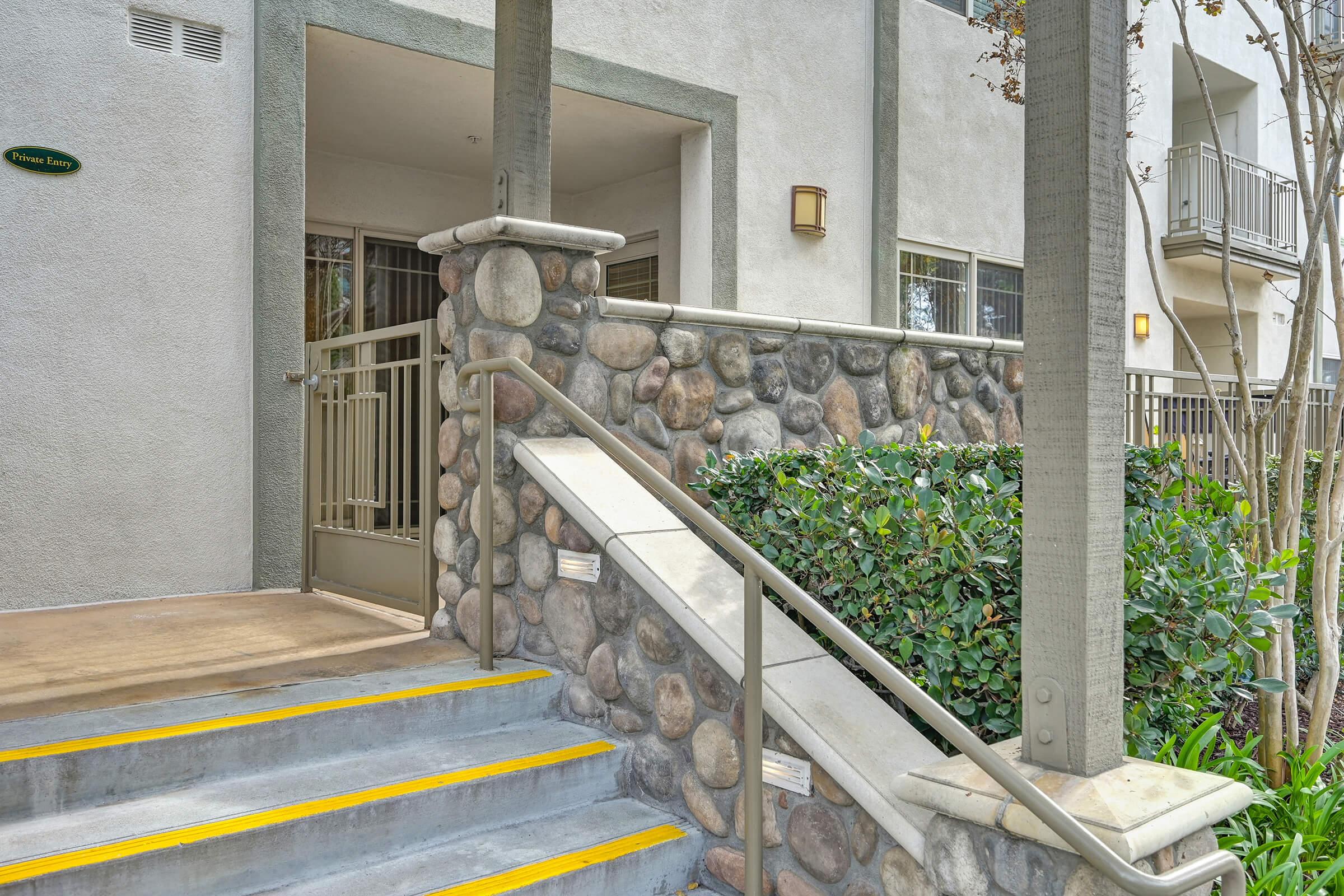 Exterior view of an apartment entrance featuring a stone wall and a staircase with yellow safety strips. Lush greenery is visible along the side, and a railing runs alongside the steps leading to a doorway. The building has a modern design, with large windows and a welcoming entrance.