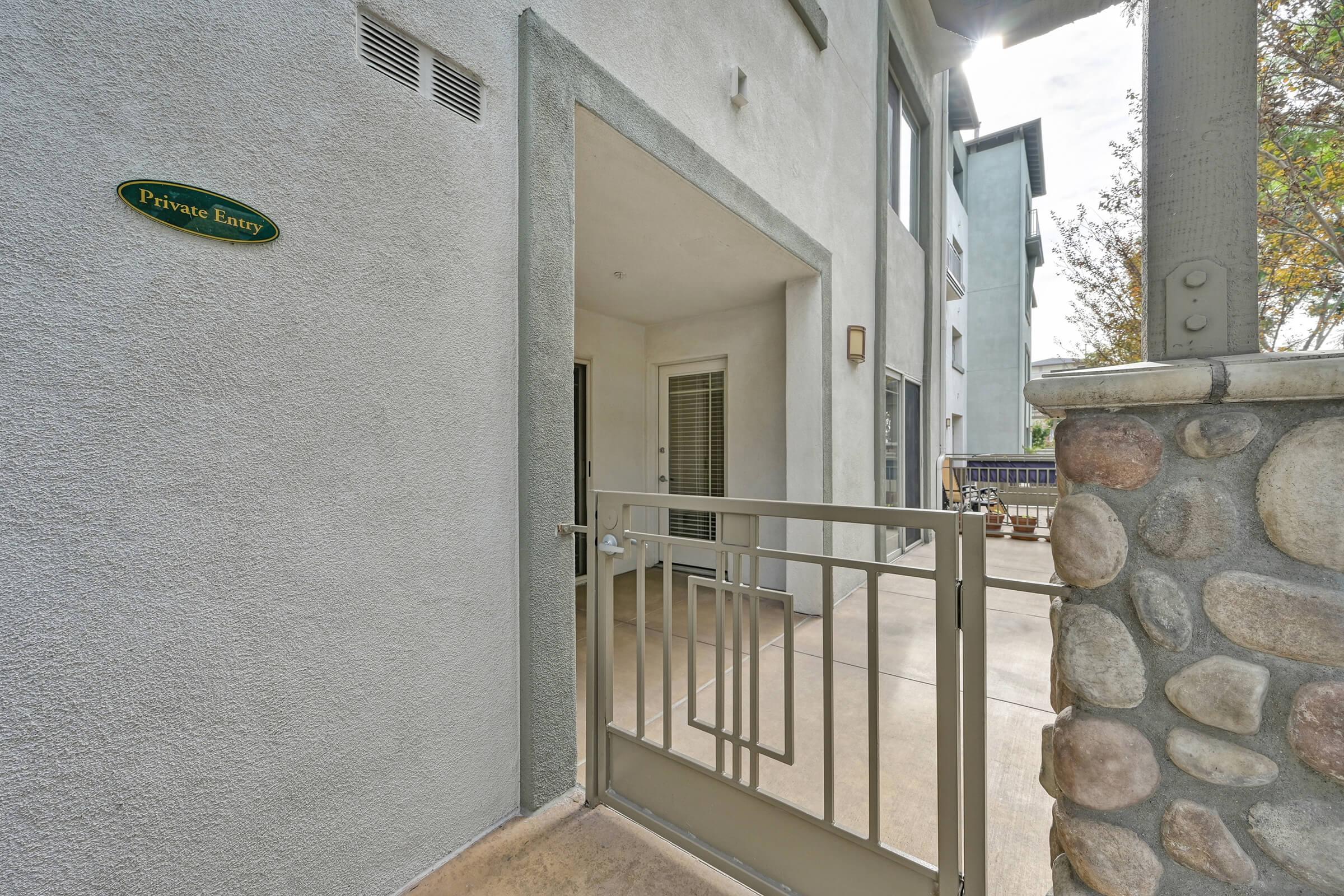A private entryway to an apartment or home, featuring a gated entrance, a stone wall, and a gray exterior. The gate is open, leading to a porch area with a concrete floor. Natural light filters in, and nearby buildings are visible in the background. A sign indicates the entrance is private.