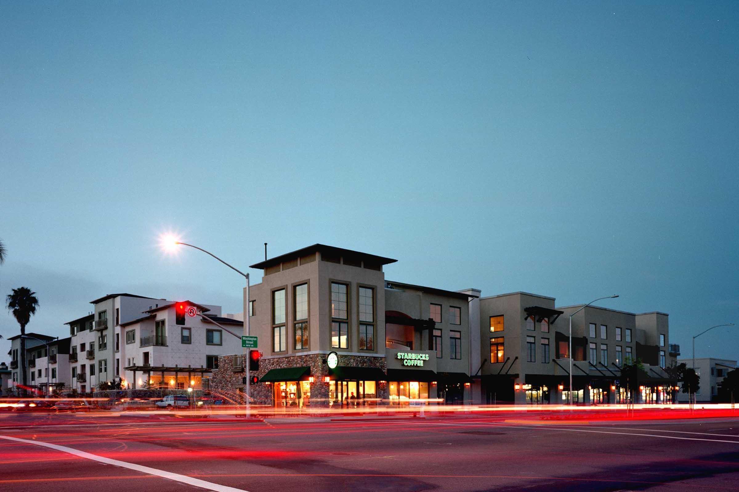 A street view at dusk featuring a modern building with a Starbucks Coffee store. The scene includes palm trees and streetlights, with light trails from passing vehicles. Nearby structures showcase contemporary architecture, creating an urban atmosphere.