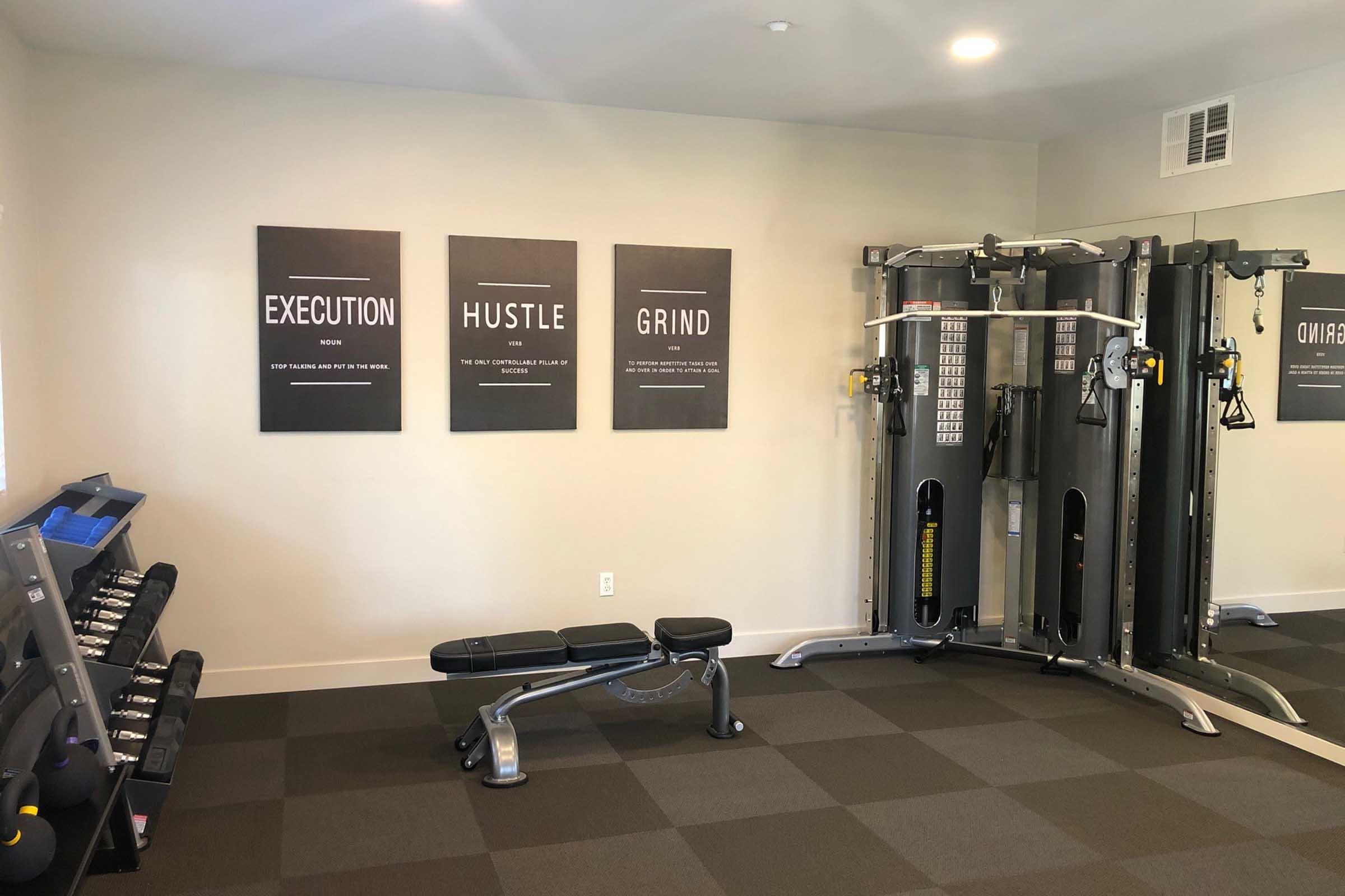 A modern gym interior featuring three motivational posters on the wall that read "EXECUTION," "HUSTLE," and "GRIND." The space includes a weightlifting bench, a cable machine, and treadmill equipment, emphasizing a fitness-oriented environment. Bright and inviting with a clean aesthetic.
